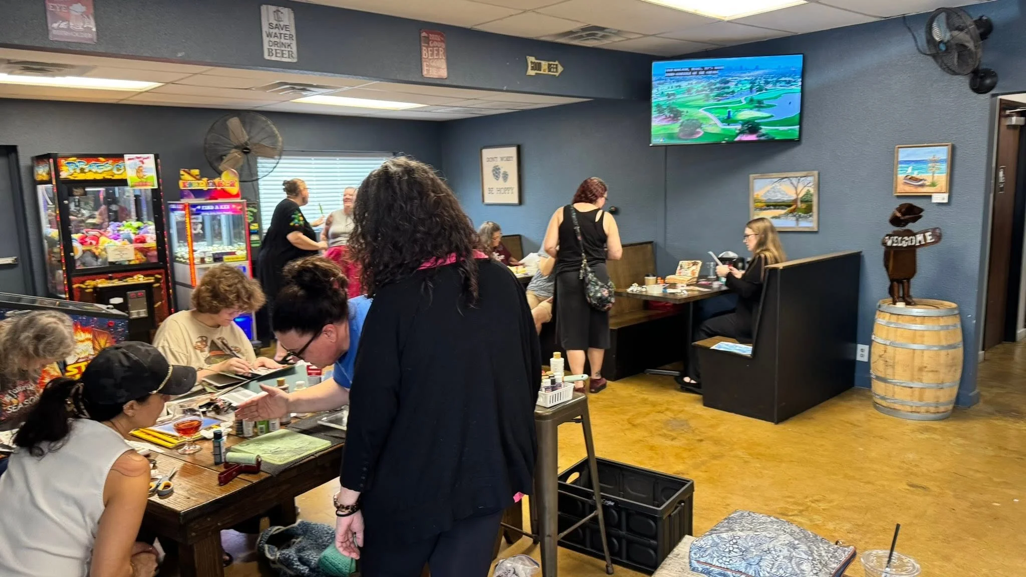 People gathered in a room engaged in arts and crafts activities with arcade games and scenic artwork on the walls.