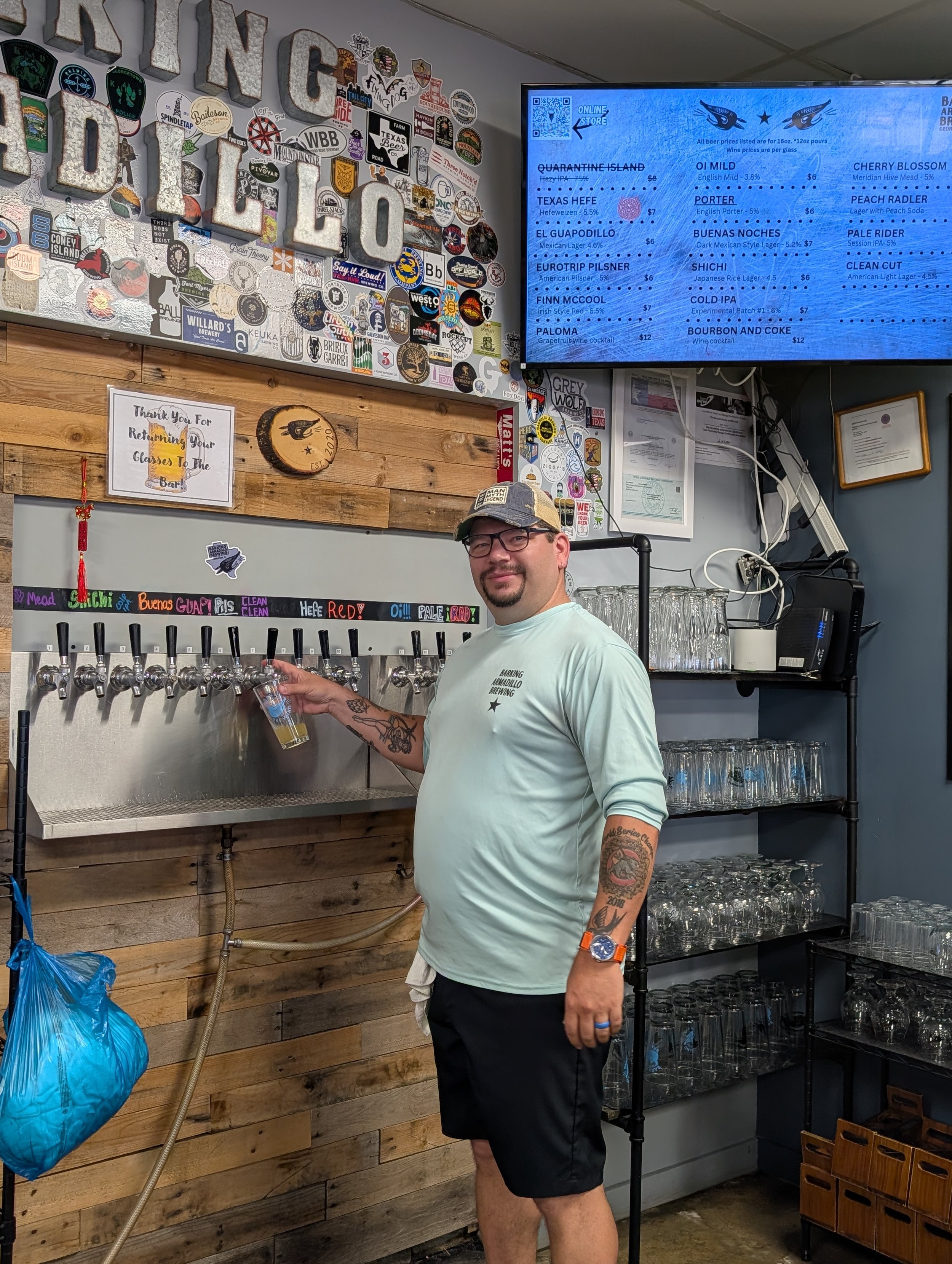 A man with glasses, tattoos, wearing a light blue T-shirt, black shorts, a baseball cap, and an orange watch, is standing in front of a beer tap handle, pouring a beer. The background features a wooden wall with various stickers and a blue digital menu board displaying different beers and prices.