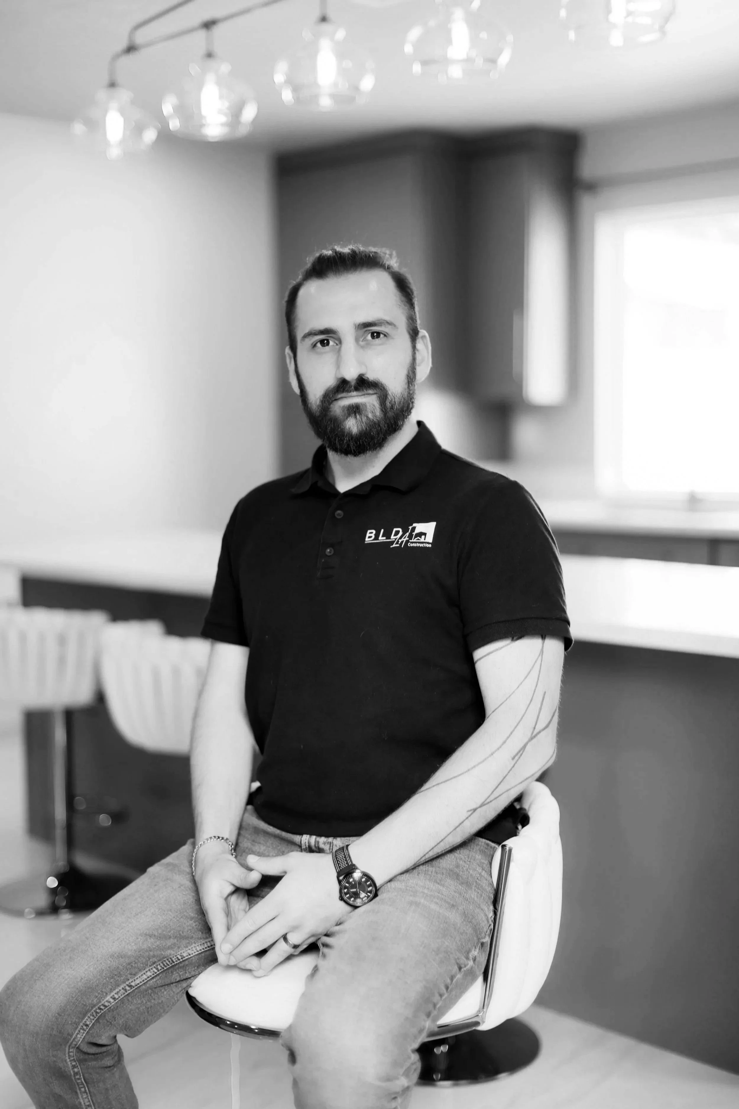 Black and white photo of a man with a beard sitting on a chair with a chain on his wrist, wearing a polo shirt and jeans, in a room with a window and ceiling lights.