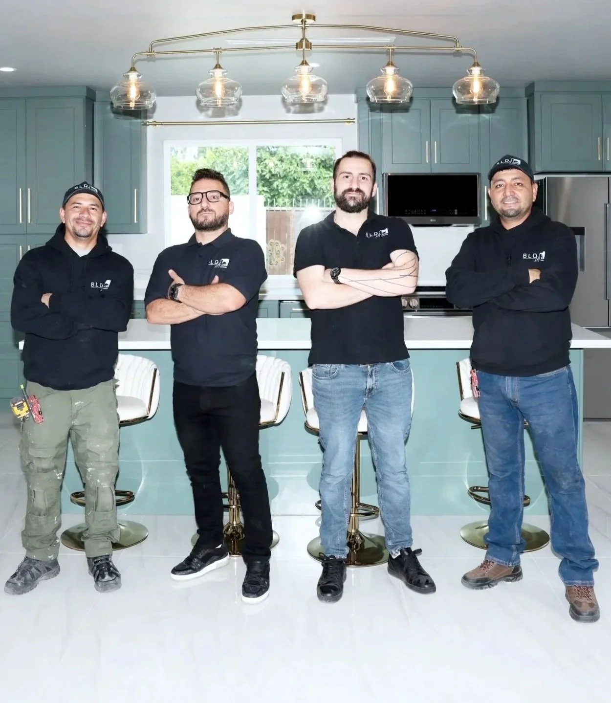 Four men standing side by side in a kitchen, all wearing black hoodies with the company logo 'BLD' on the chest, arms crossed, with modern green cabinets and a window behind them.
