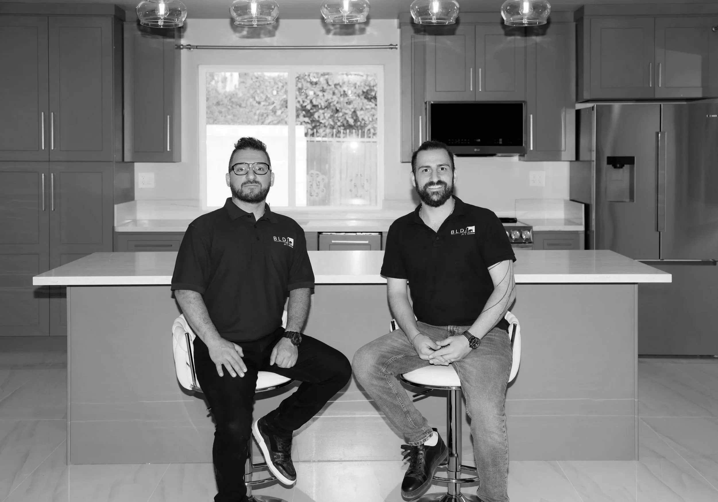Two men sitting in kitchen with gray cabinets and window in background