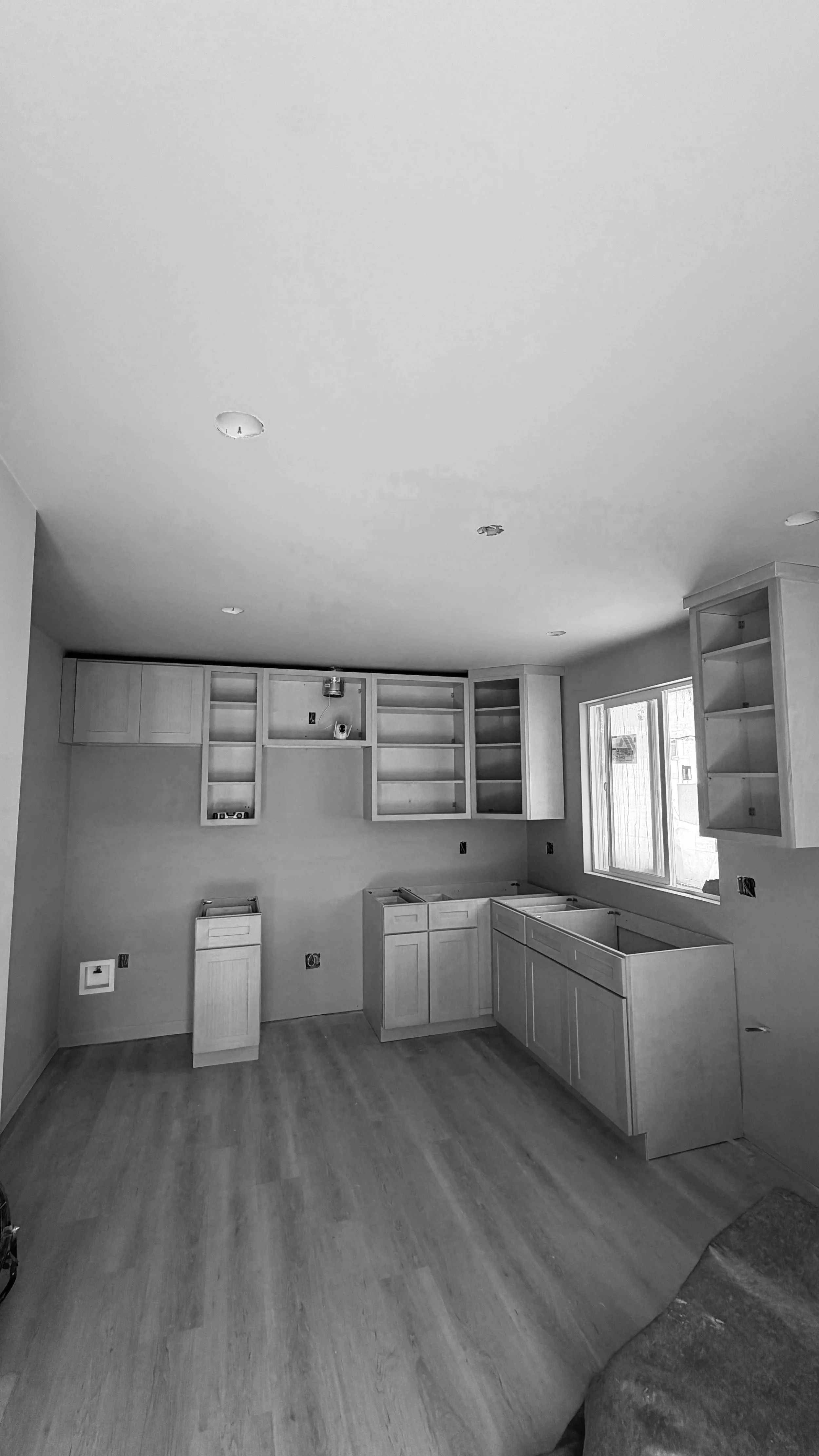 Kitchen under construction with light-colored cabinets, a window, and hardwood floors, with electrical outlets and light fixtures not yet installed.