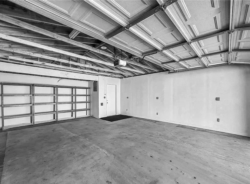 Empty residential garage with concrete floor, drywall walls, and an unfinished ceiling showing exposed beams and garage door mechanism.