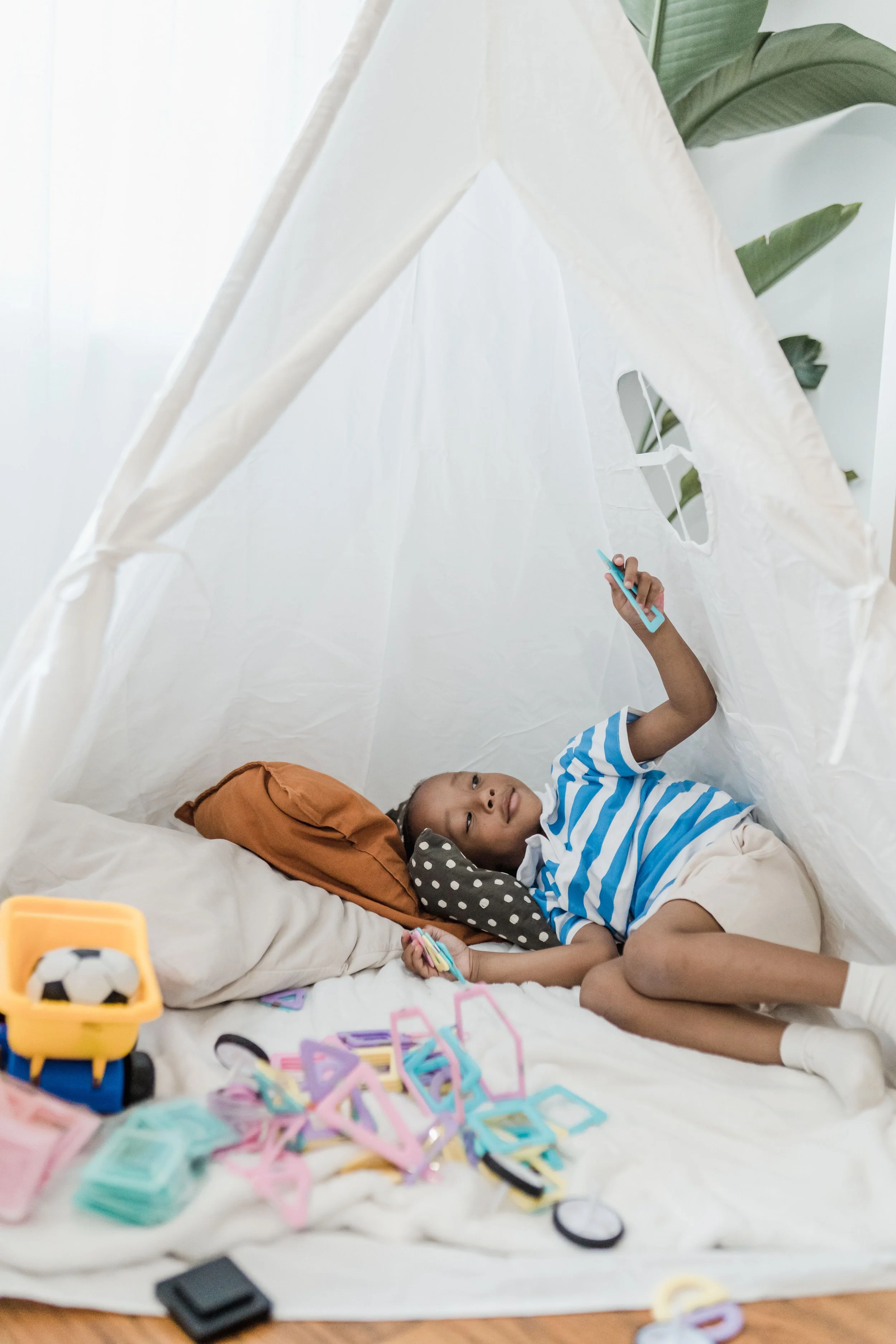A boy lying inside a white fabric play tent, holding a toy phone, surrounded by colorful plastic building blocks and toys, with some leaves visible outside the tent.