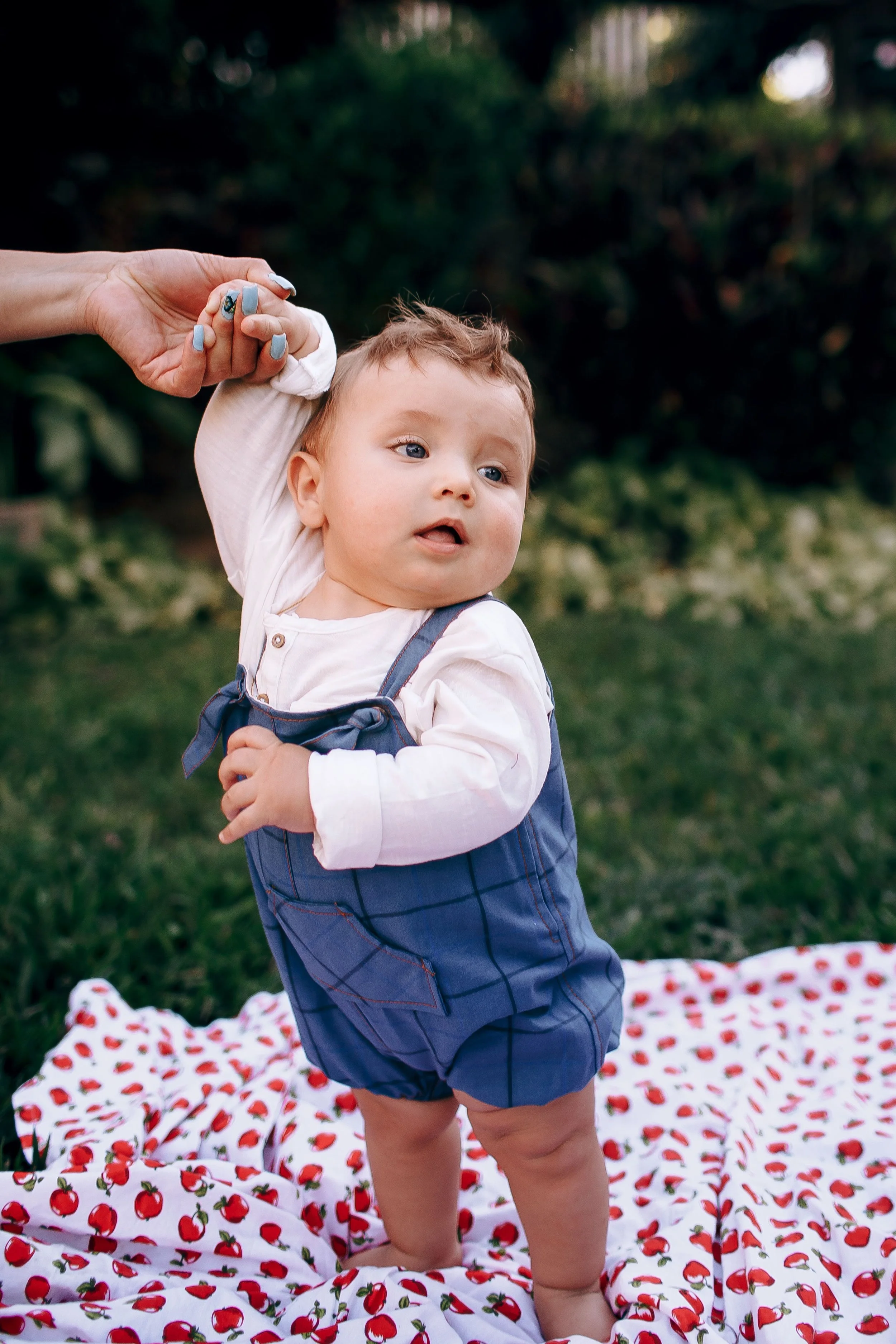 A young child with curly hair and blue eyes standing on a cherry-patterned blanket outdoors, holding an adult's hand for support as they walk.