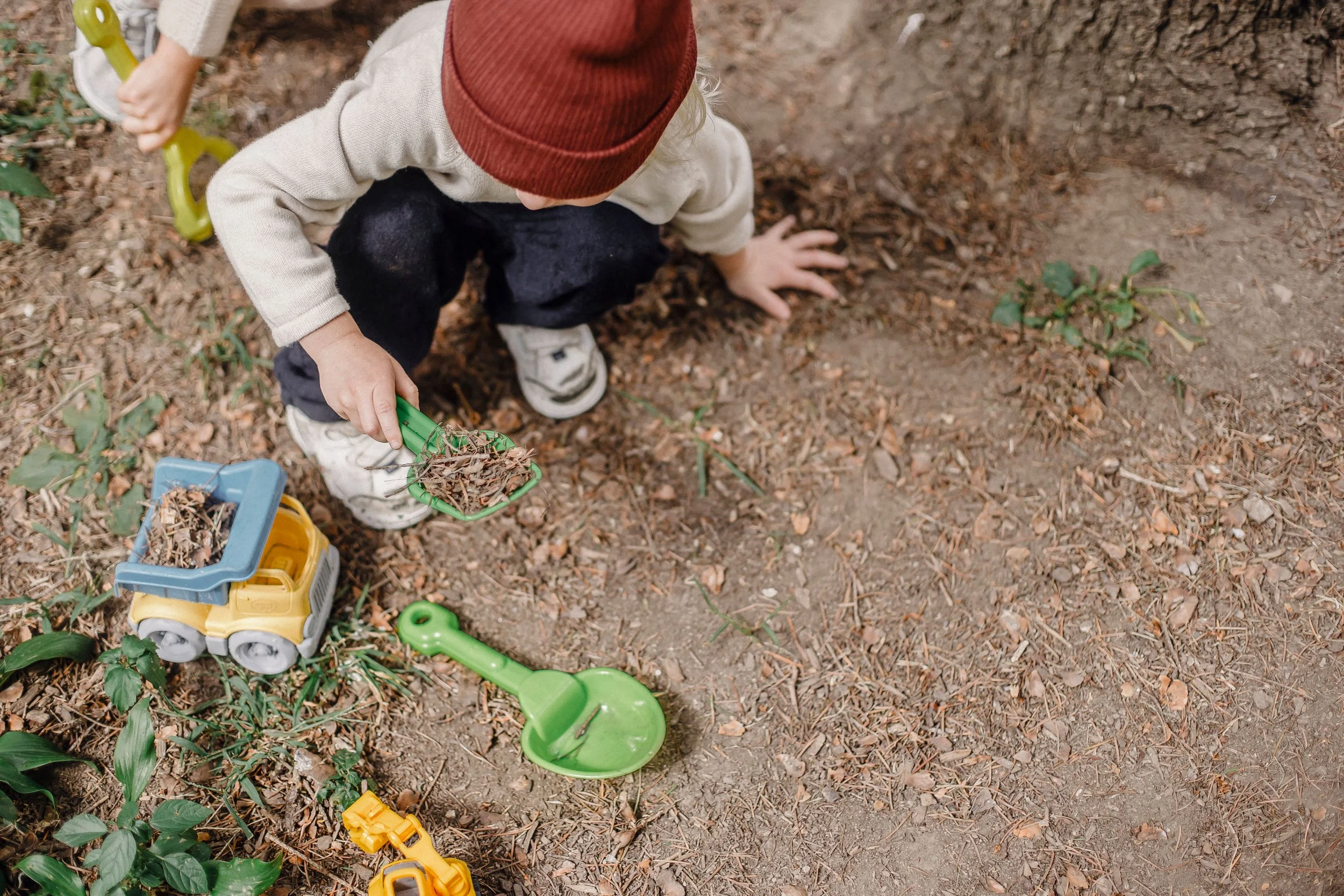Child crouching on dirt ground, scooping leaves into a small green toy shovel, with toy trucks and gardening tools scattered around.