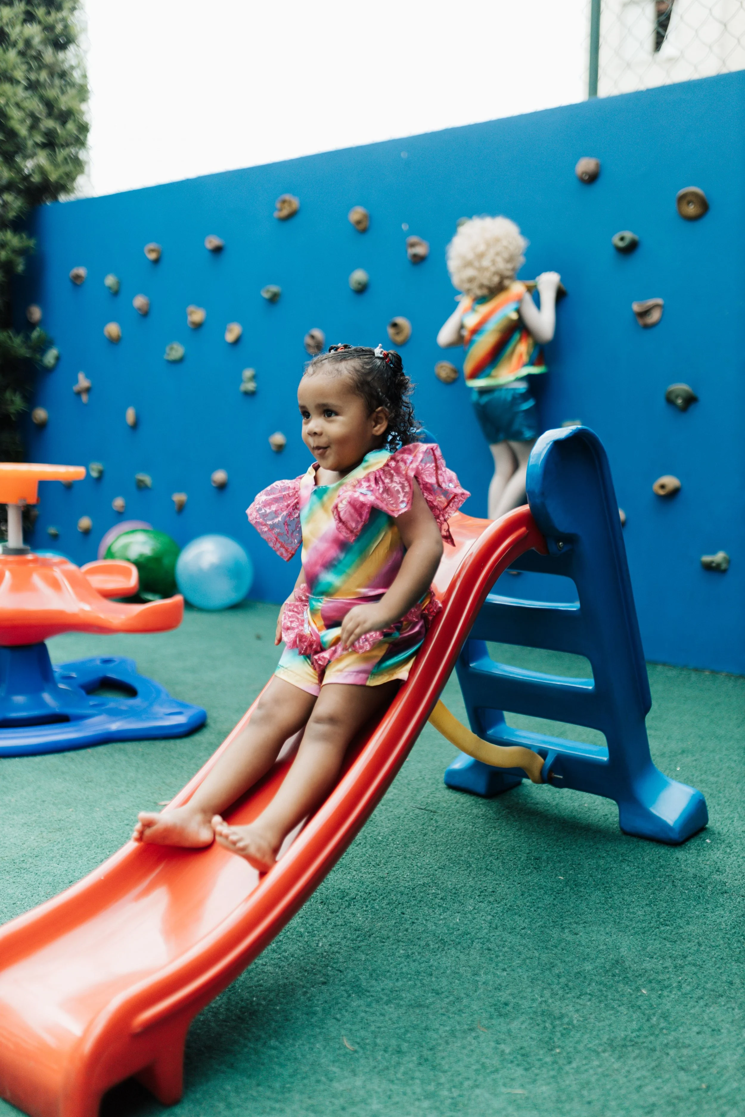 Two children playing on a slide in a playground. One girl with curly hair sits at the top of the orange slide, smiling. The other child, with an afro hairstyle, climbs a blue climbing wall in the background.