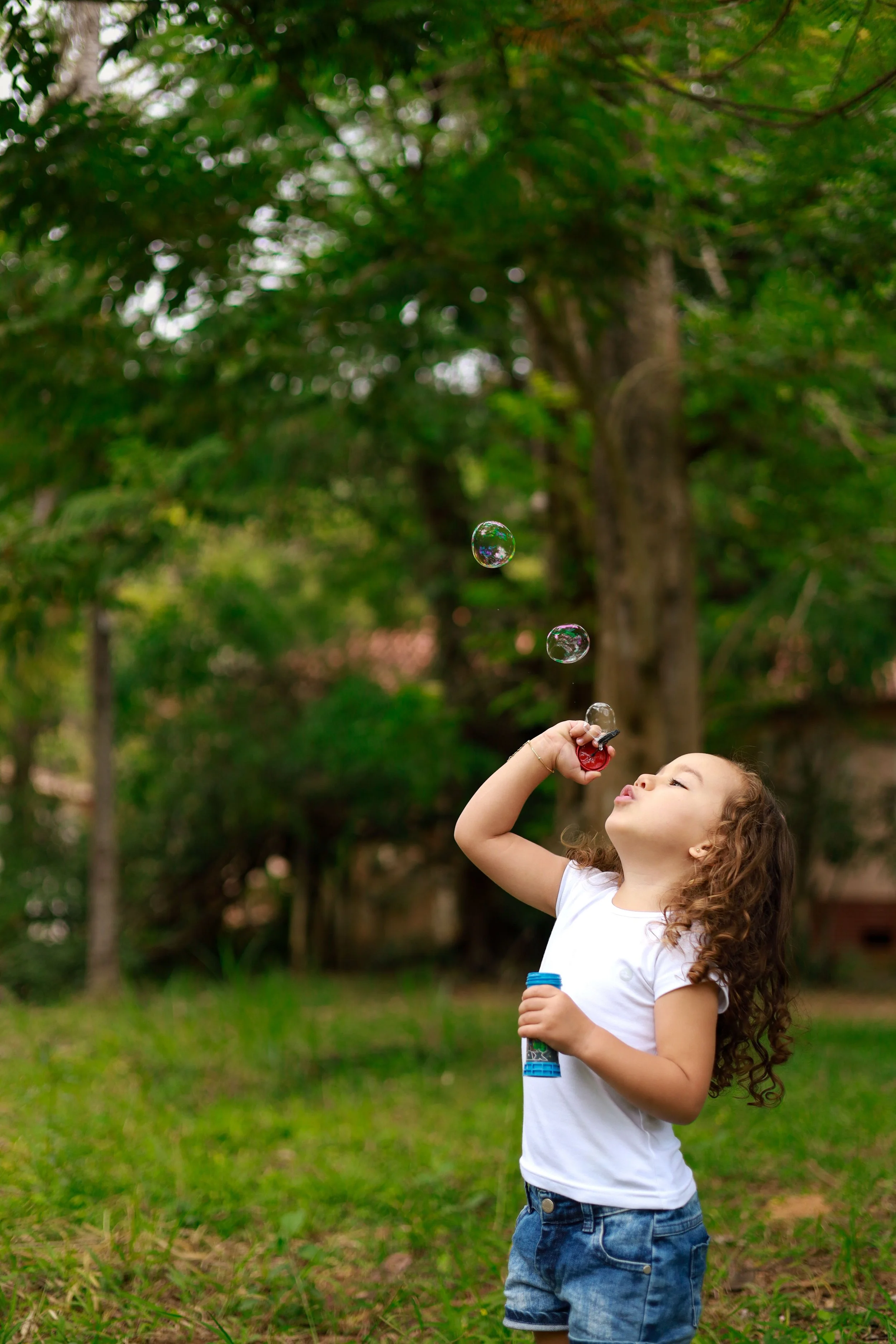 Young girl with curly hair wearing a white T-shirt and denim shorts, playing with soap bubbles outdoors in a green park.