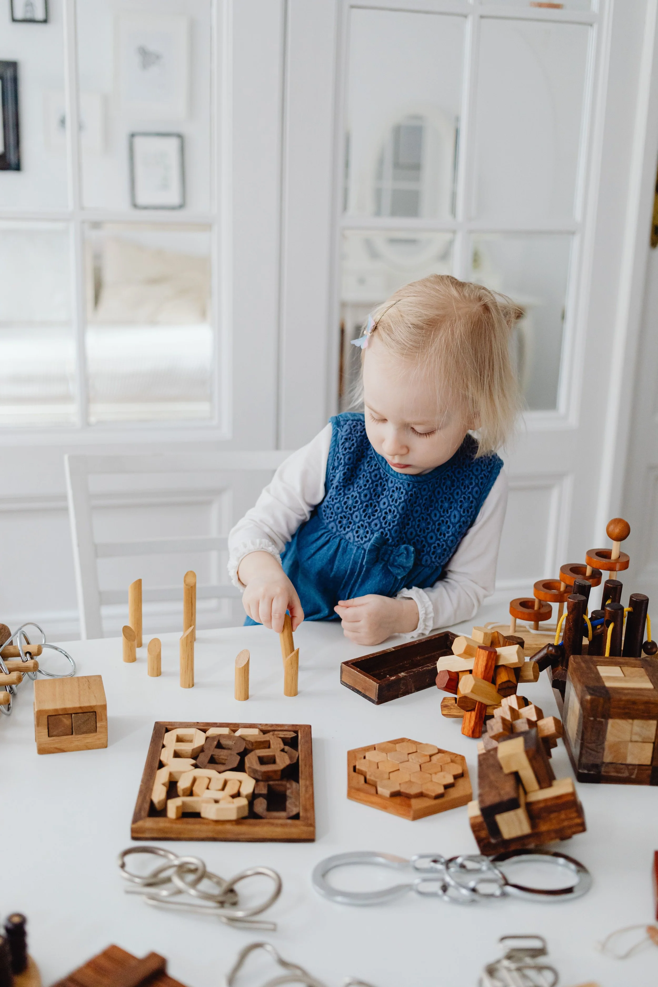 A young girl with blonde hair wearing a blue dress and white long-sleeve shirt playing with wooden toys on a white table in a bright room.