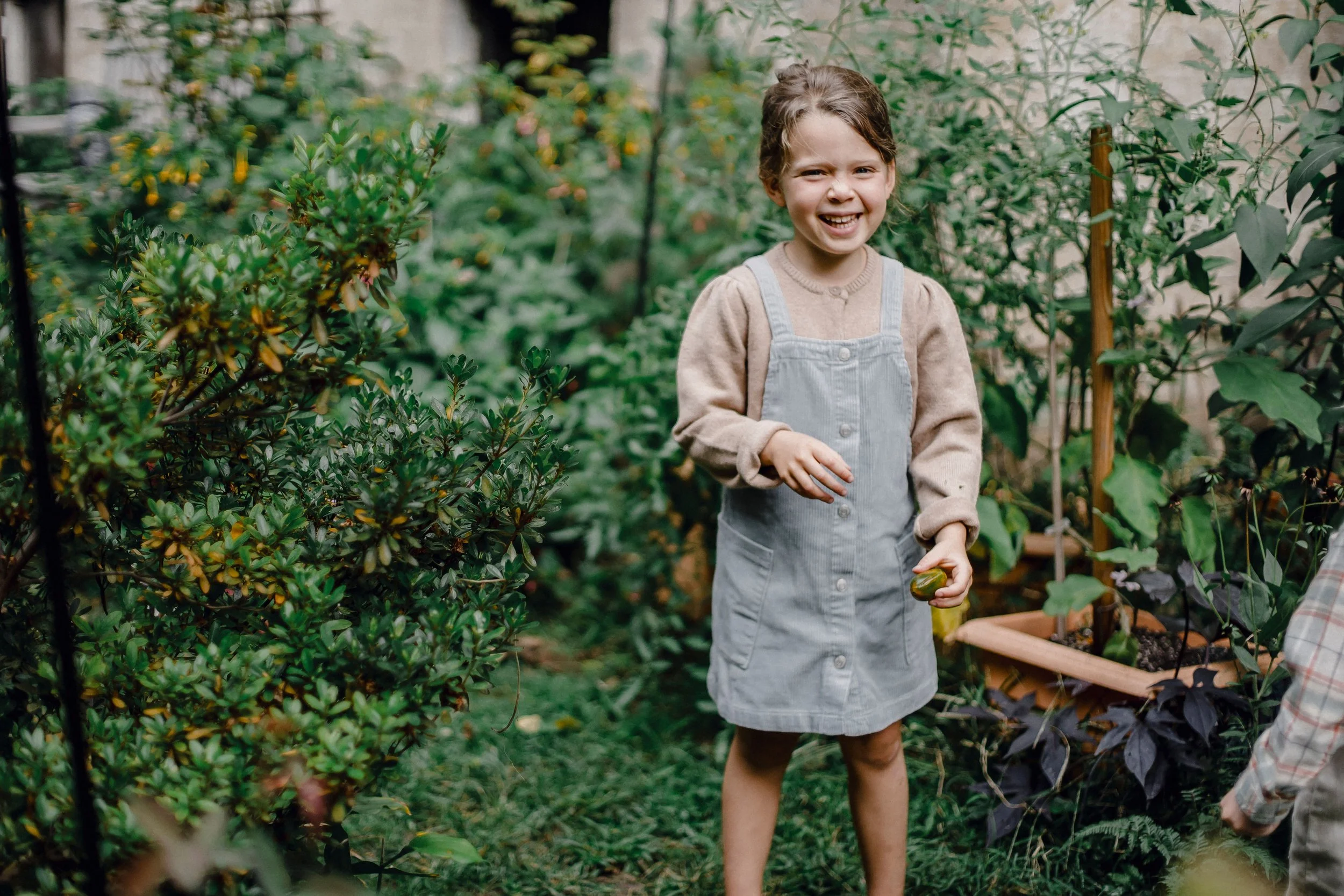 A young girl with short brown hair smiling and wearing a beige sweater and gray overalls, standing in a lush backyard garden surrounded by green plants and flowers.