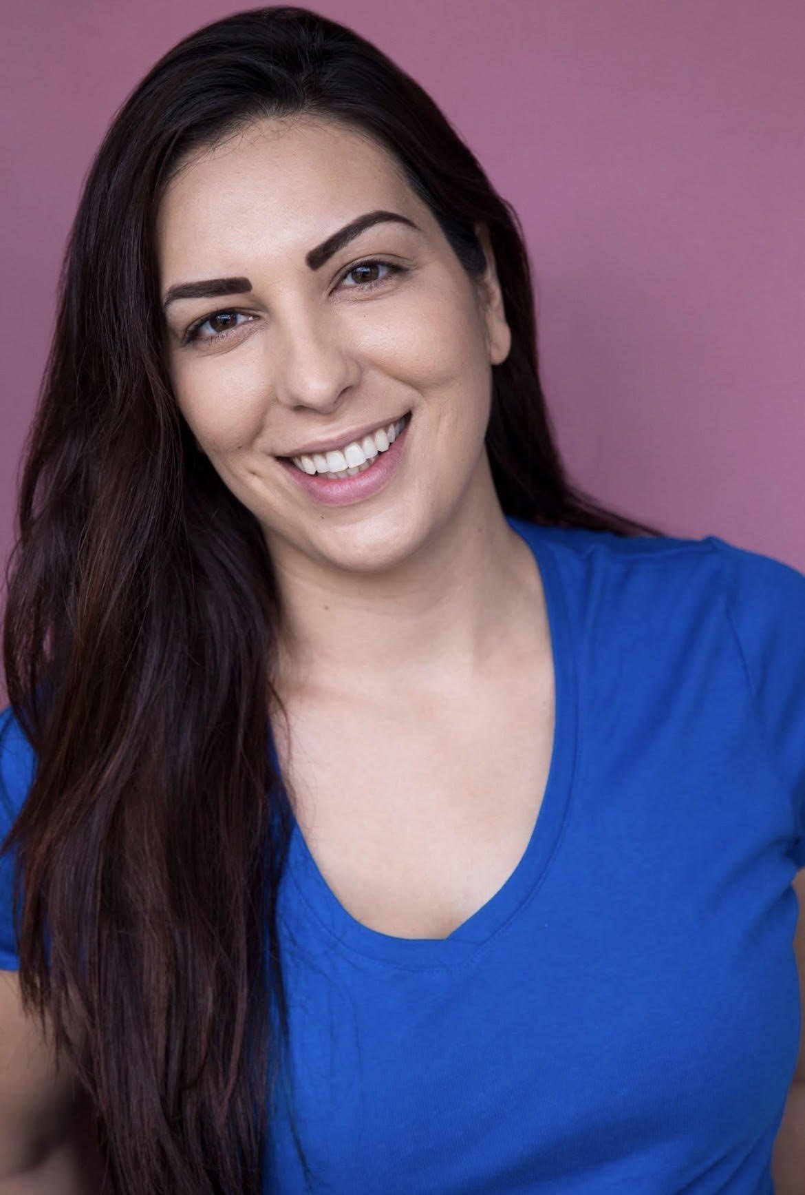 A woman with long dark hair smiling against a pink background, wearing a blue top.