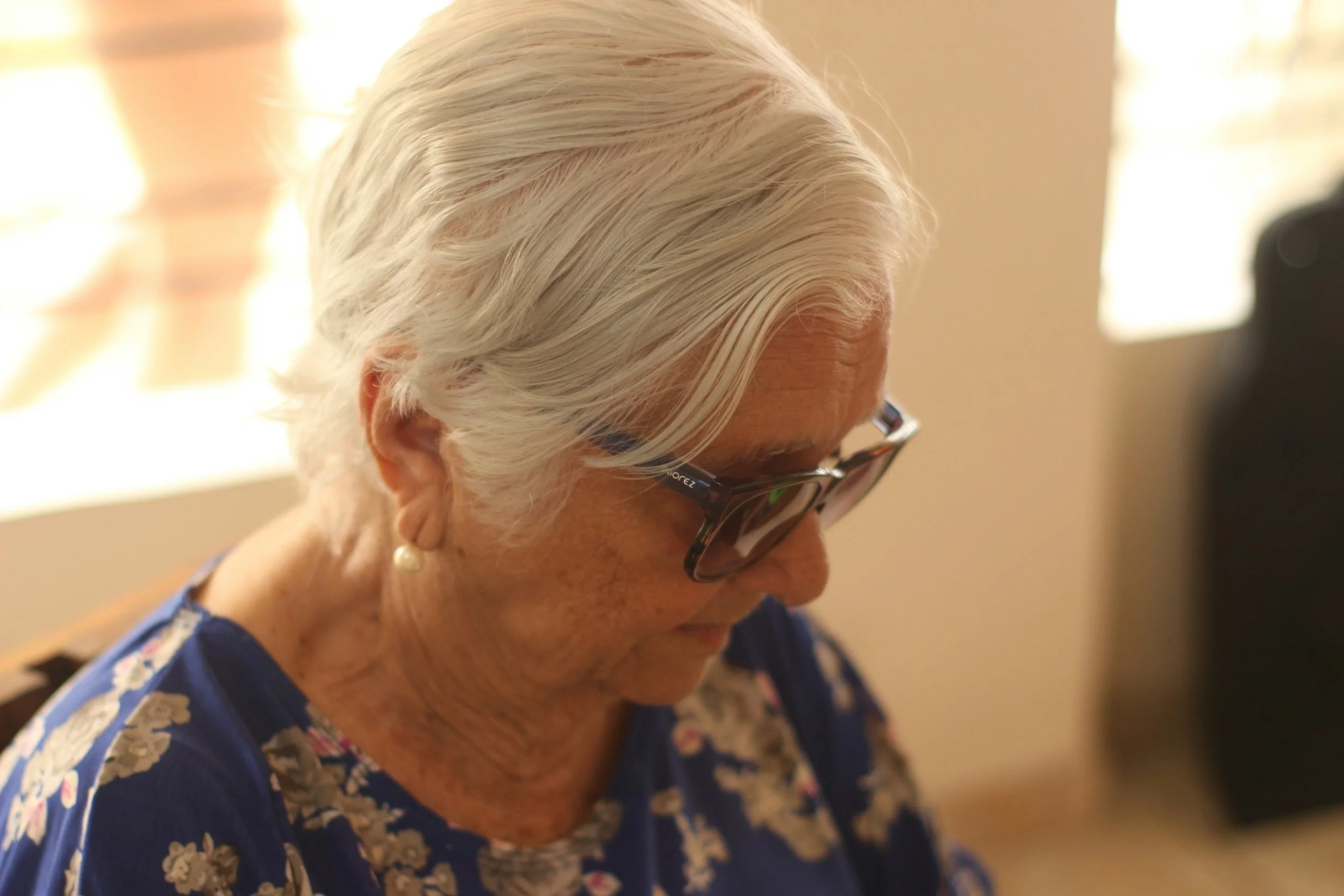 Close-up of an elderly woman with white hair wearing glasses, a floral top, and pearl earrings, indoors with soft lighting.