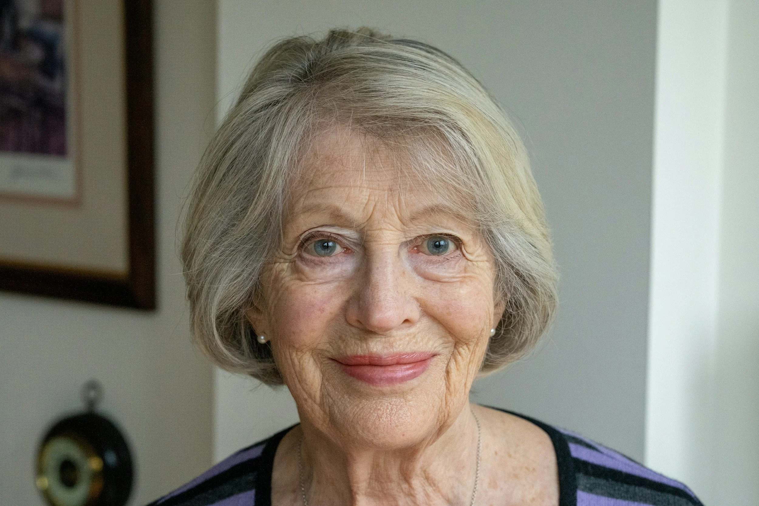 Close-up of an elderly woman with short gray hair, blue eyes, and earrings, smiling at the camera.