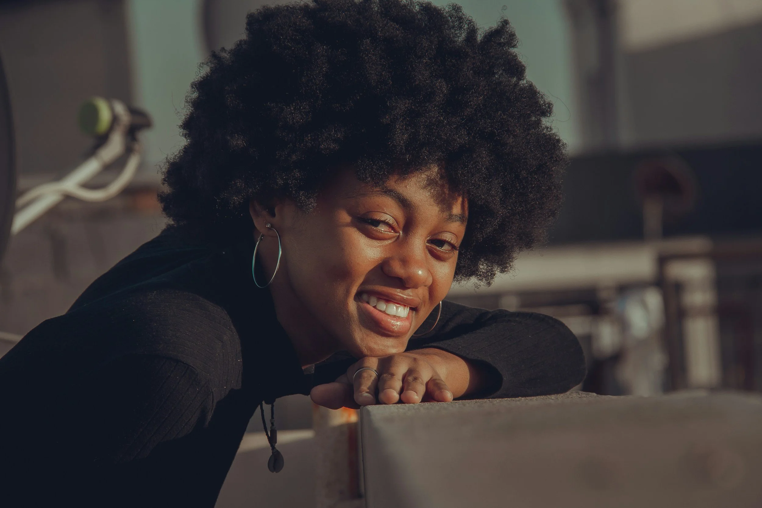 A woman with a large afro hairstyle smiling and leaning on a surface outdoors, with a blurred background of a building and a large window.