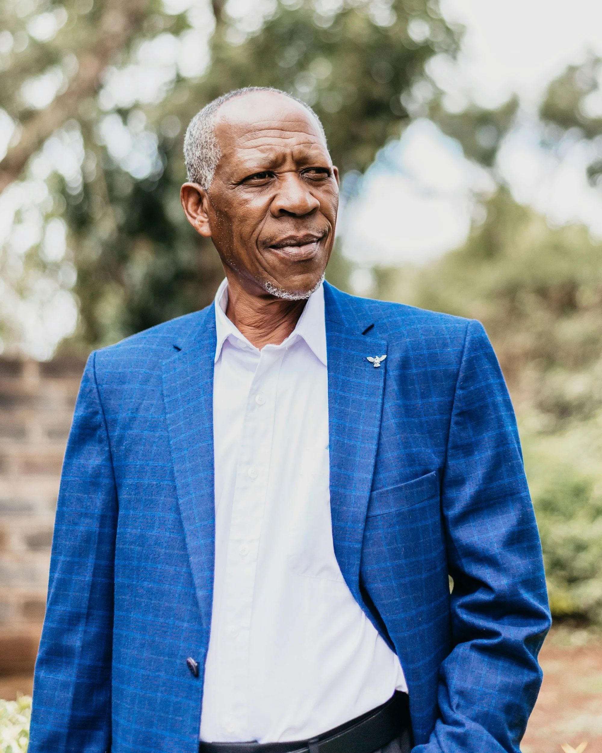 Close-up of an elderly man wearing a blue blazer with a small eagle pin on the lapel, white shirt, and black belt, outdoors with blurred trees in the background.