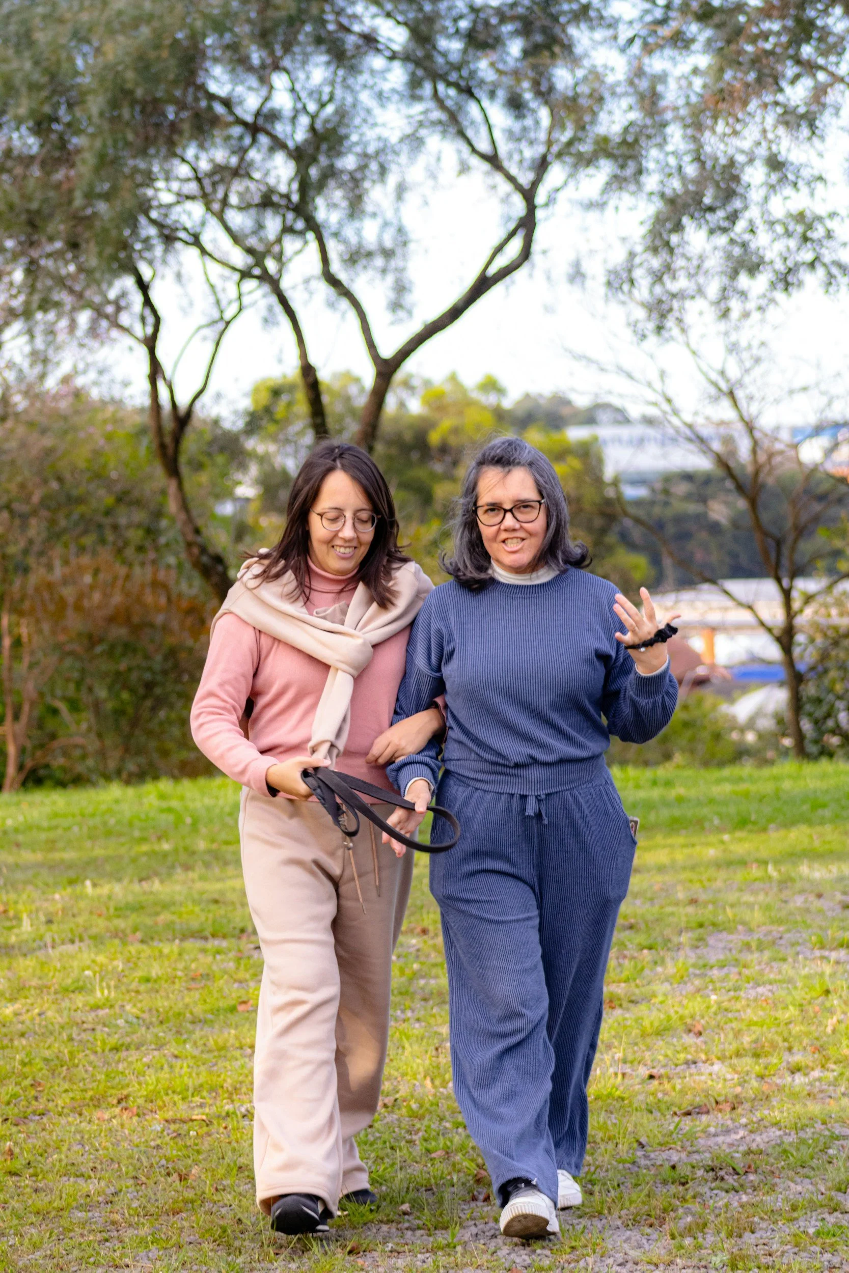 Two women walking in a park, smiling and talking, with trees and buildings in the background.