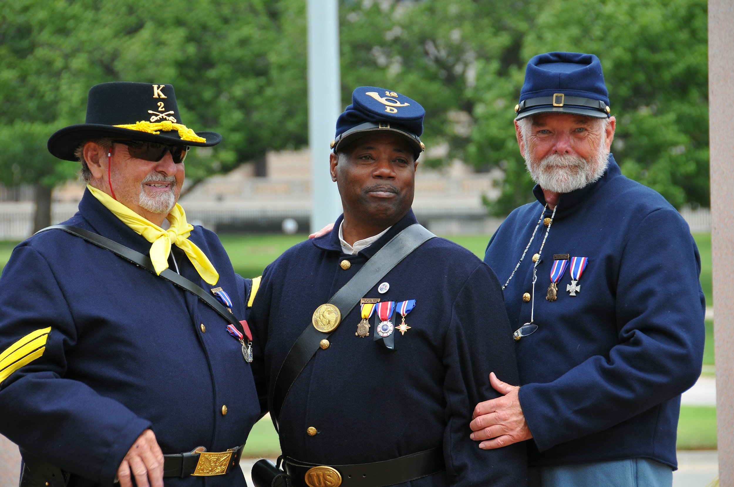Three older men in vintage military uniforms pose together outdoors, wearing hats with medals and decorations, with trees in the background.