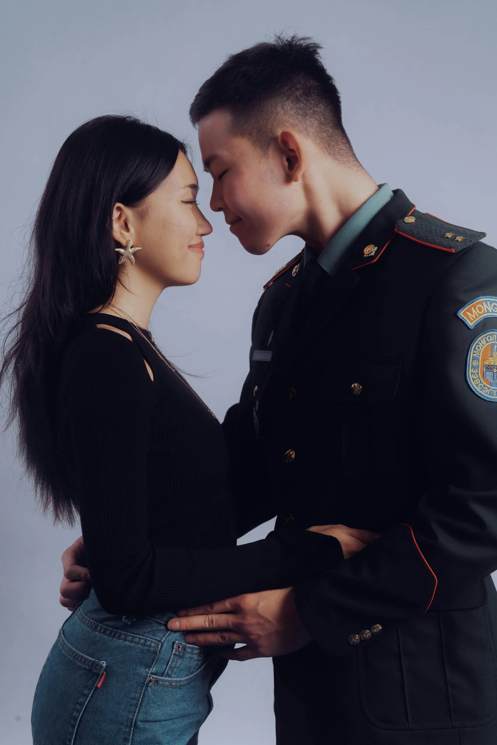 A young woman and a young man are standing close with their foreheads touching, smiling softly, and holding each other by the waist and hips. The woman has long dark hair, star-shaped earrings, and is wearing a black top and jeans. The man is in a dark police uniform with badges and patches, including a patch that says 'MONG' and other insignia. The background is plain and gray.