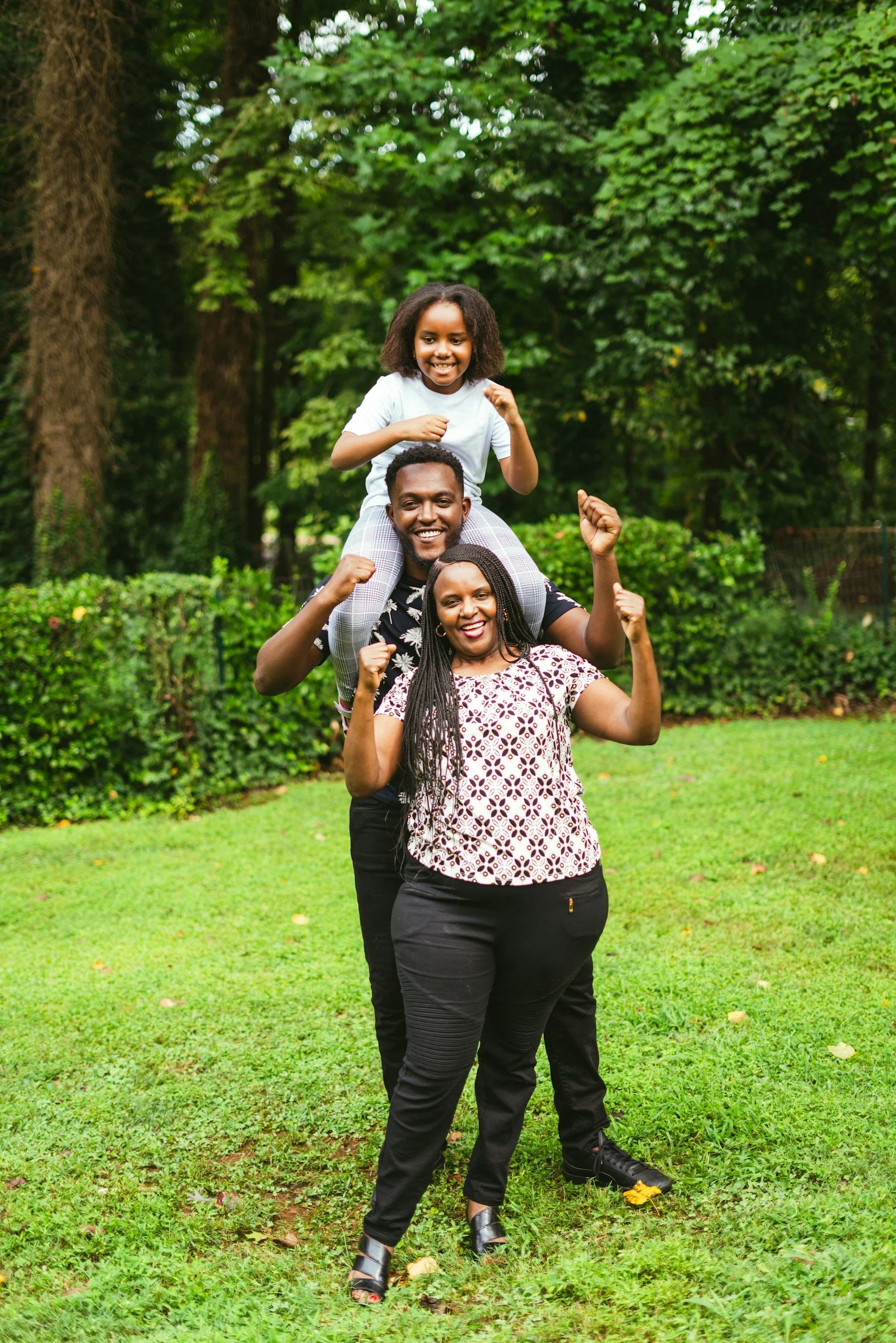 A family of three celebrating outdoors in a park, with the woman standing in front, the man behind her with his arms raised, and a girl sitting on his shoulders, all smiling and showing raised fists.