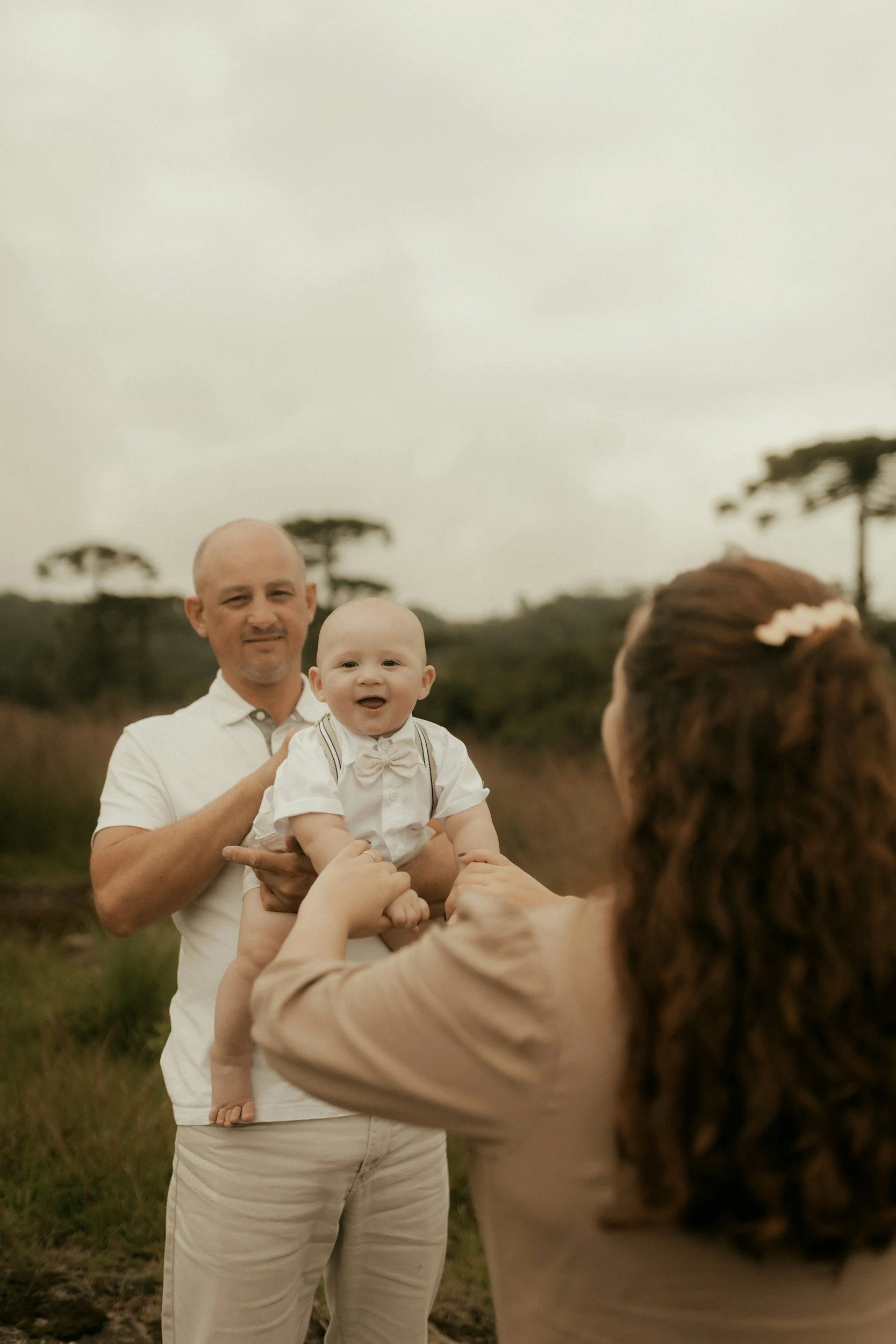 A man holding a smiling baby boy outdoors, with a woman in the foreground reaching towards the baby, in a natural landscape with trees in the background.