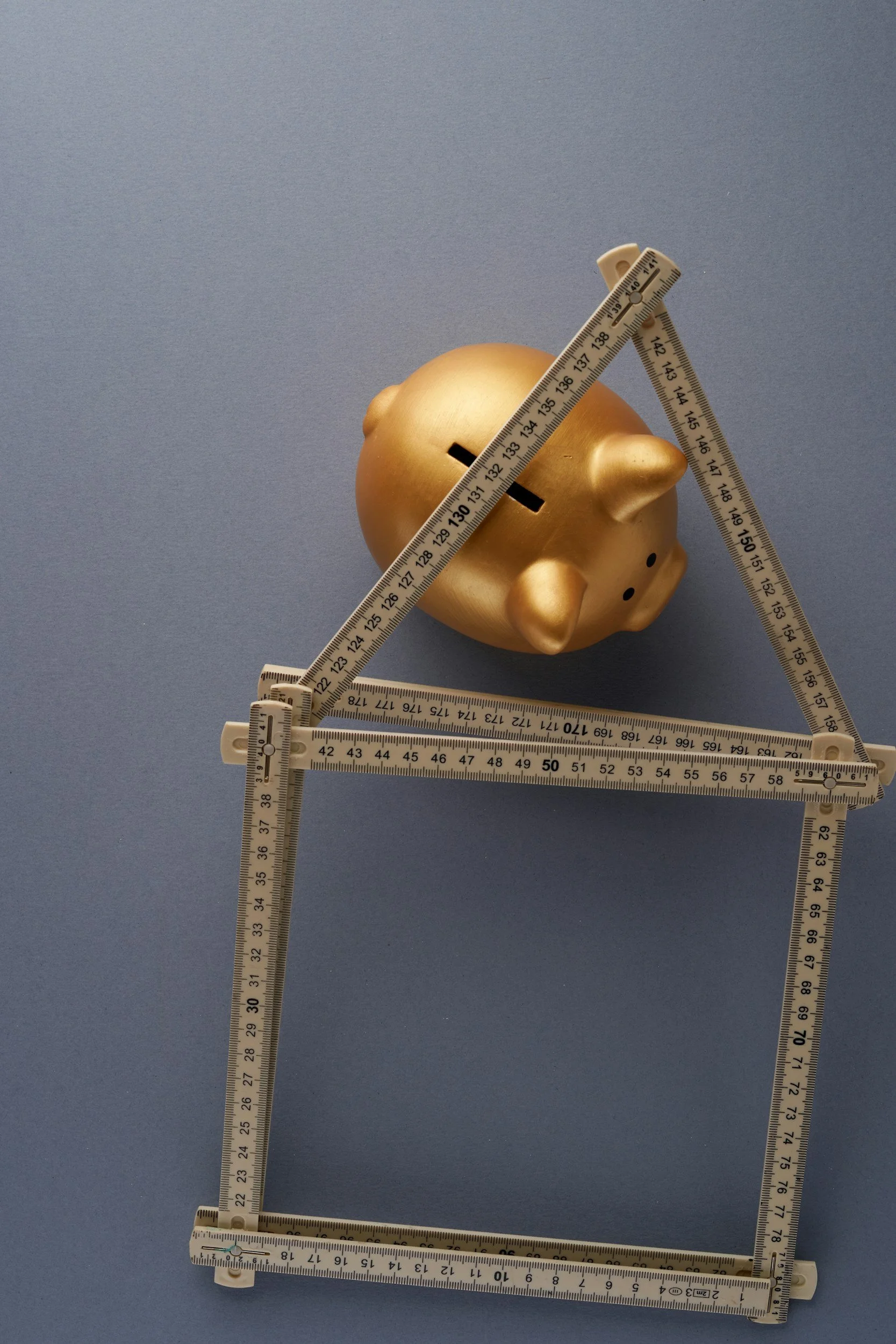 A gold piggy bank with a slot on its back, surrounded by folding white rulers with black markings, on a blue background.