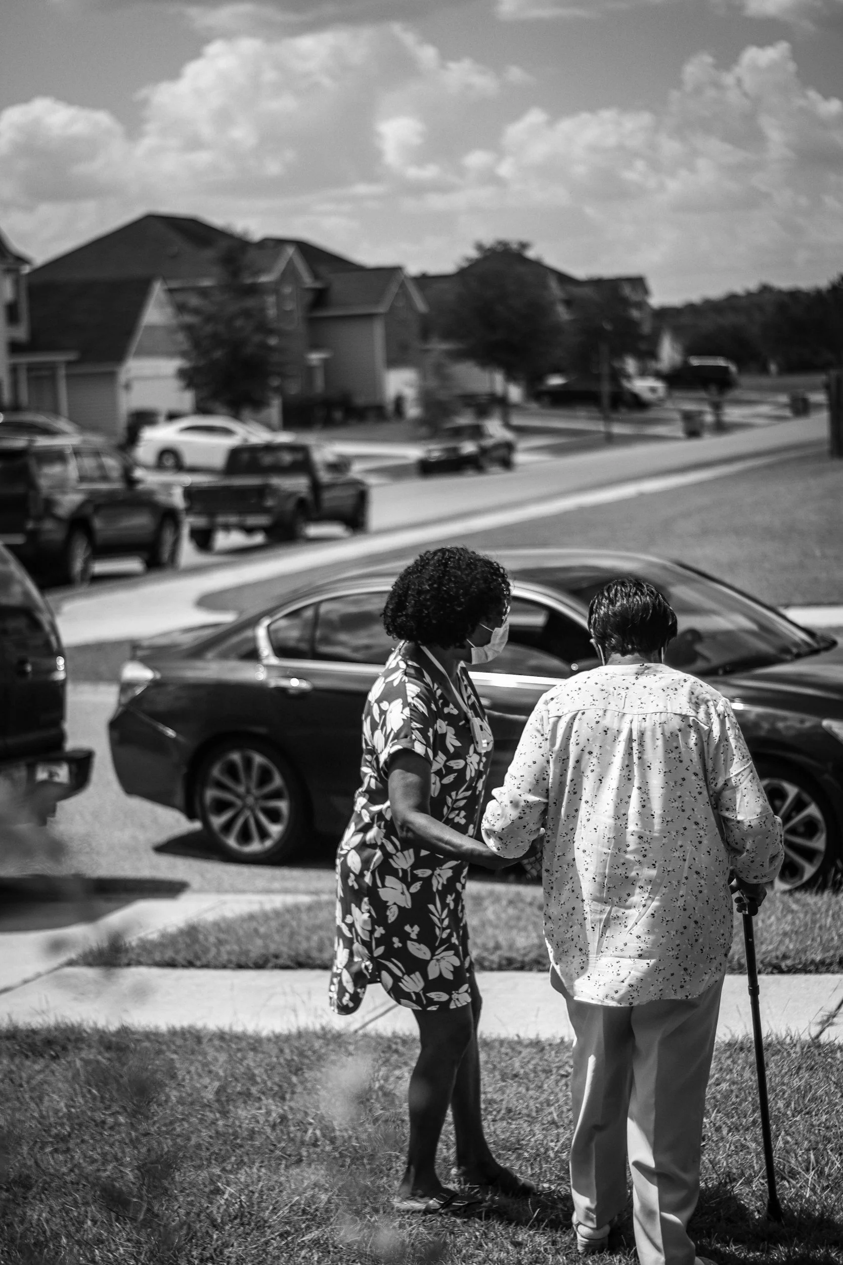 Two women walking together on a sunny day near parked cars and suburban houses, one wearing a face mask and the other using a cane.