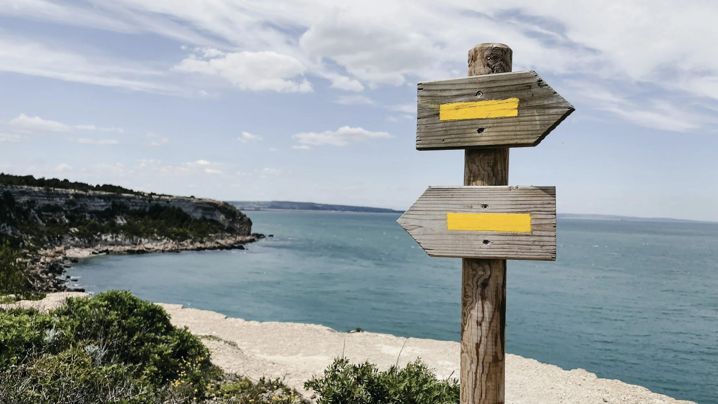 A wooden signpost with two directional arrows painted with yellow stripes, situated near a coastal area with cliffs and the ocean in the background under a partly cloudy sky.