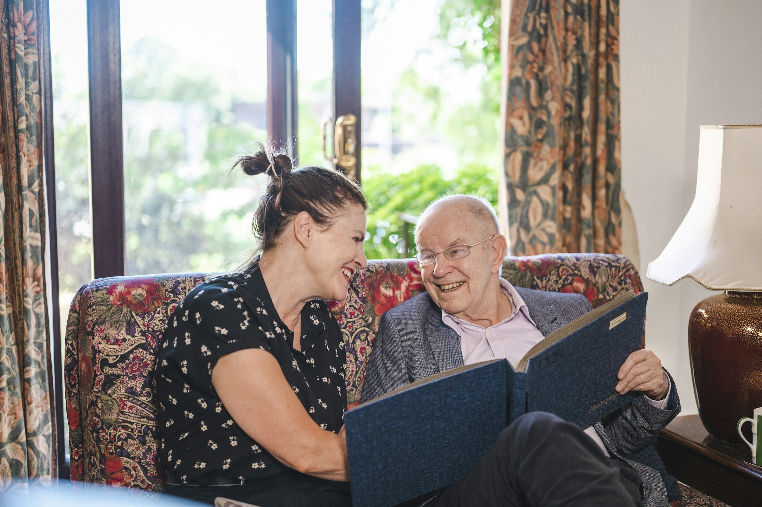Two women and an elderly man sitting together on a floral couch, looking at a photo album with smiles, near large glass doors with curtains and greenery outside.