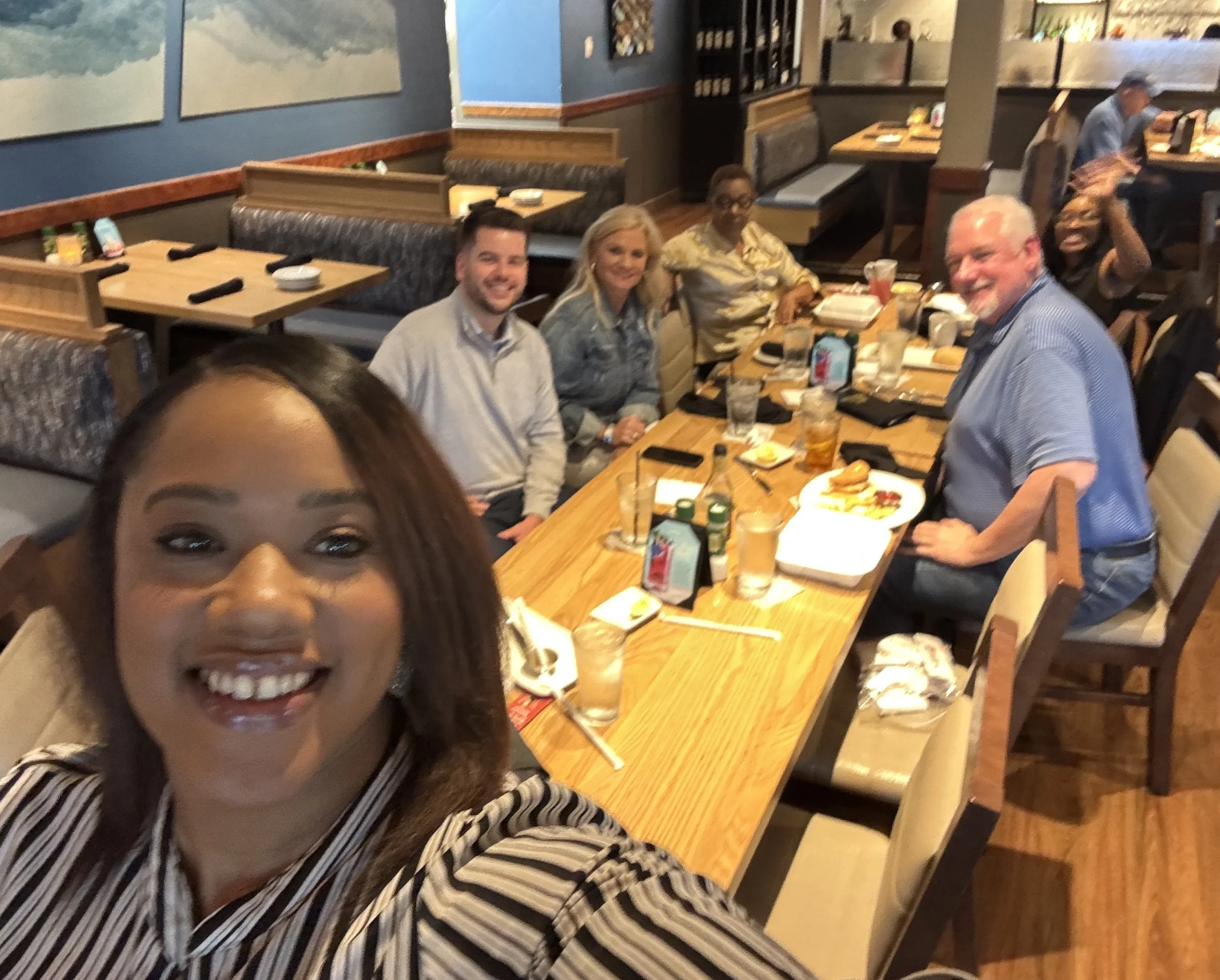 Group of people sitting around a restaurant table, smiling and enjoying food and drinks, with a woman taking a selfie in the foreground.