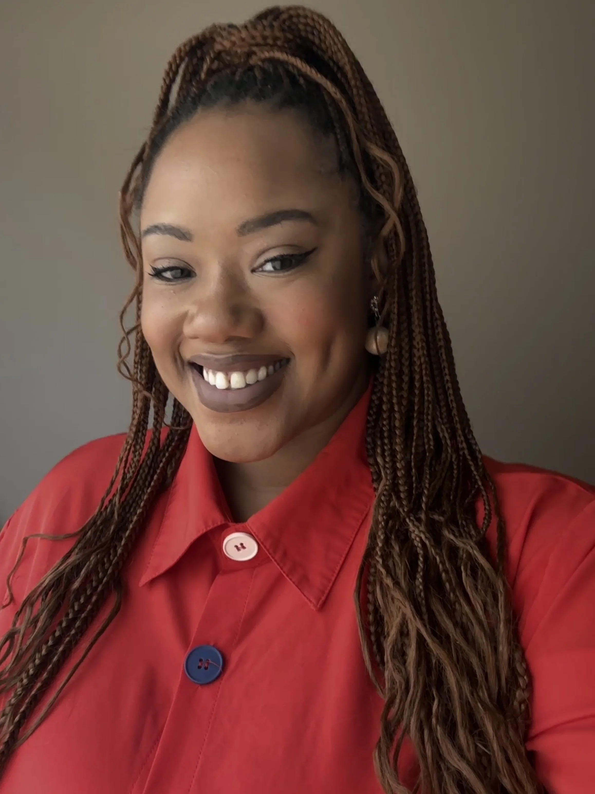 A woman with long braided hair and dark lipstick, smiling and wearing a red jacket with blue and white buttons.