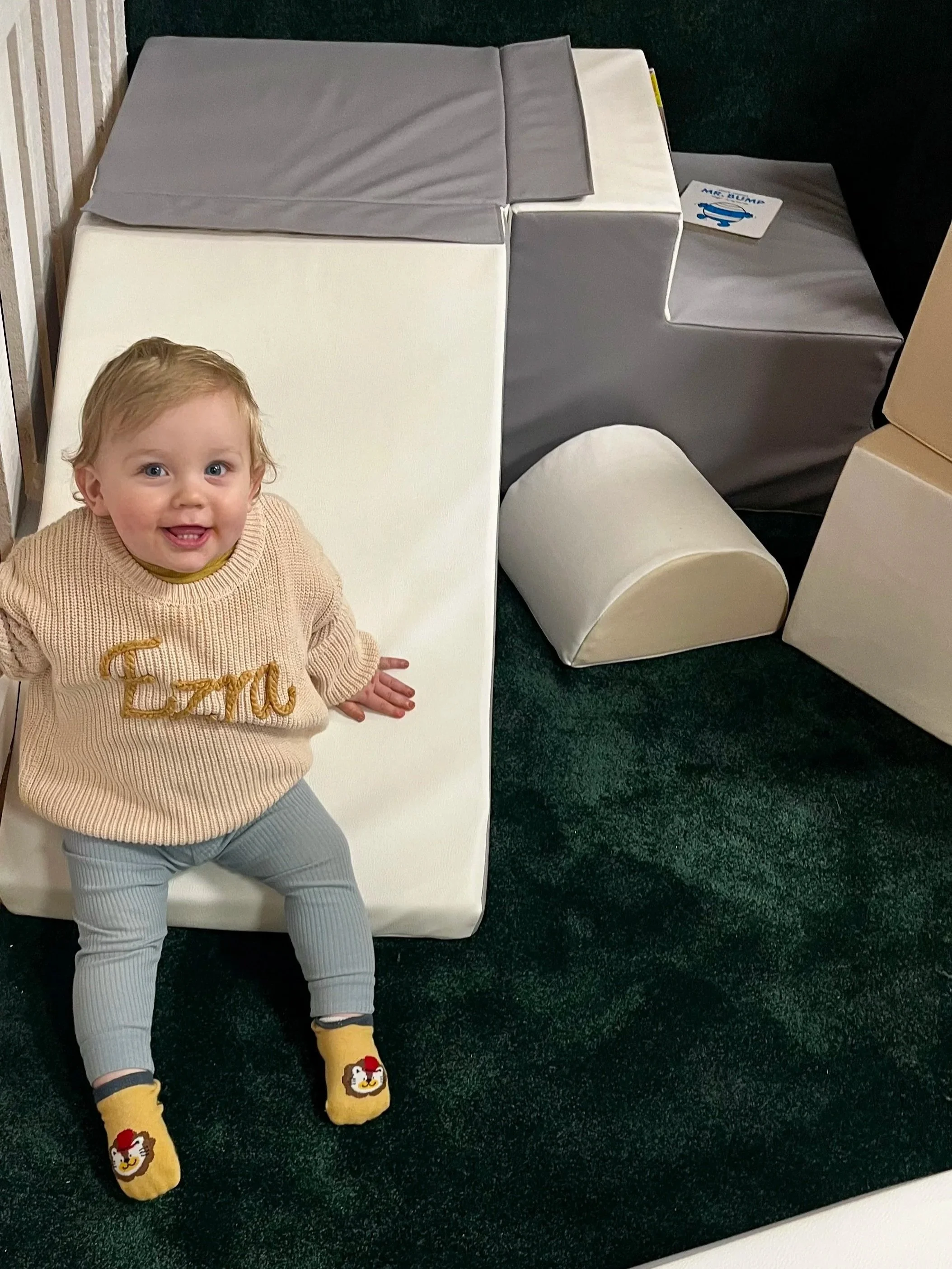 A young child with curly blonde hair and blue eyes is sitting on a white foam padded surface, smiling and looking up. The child is wearing a beige sweater with the word 'Dream' embroidered in gold on the front, gray pants, and yellow socks with cartoon lion faces.