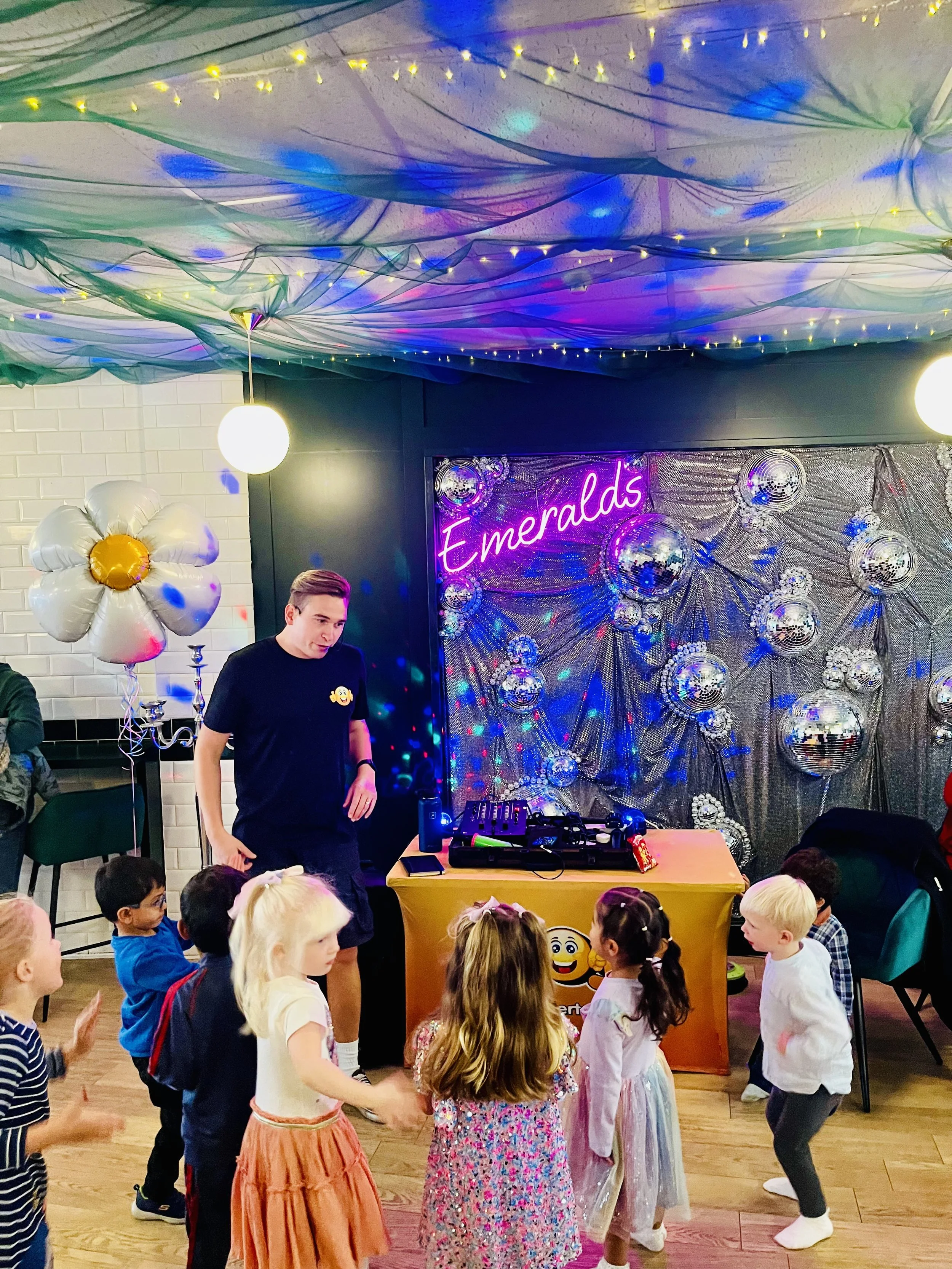 Children dancing at a birthday party with a DJ, colorful decorations, and a neon sign that reads 'Emeralds'.