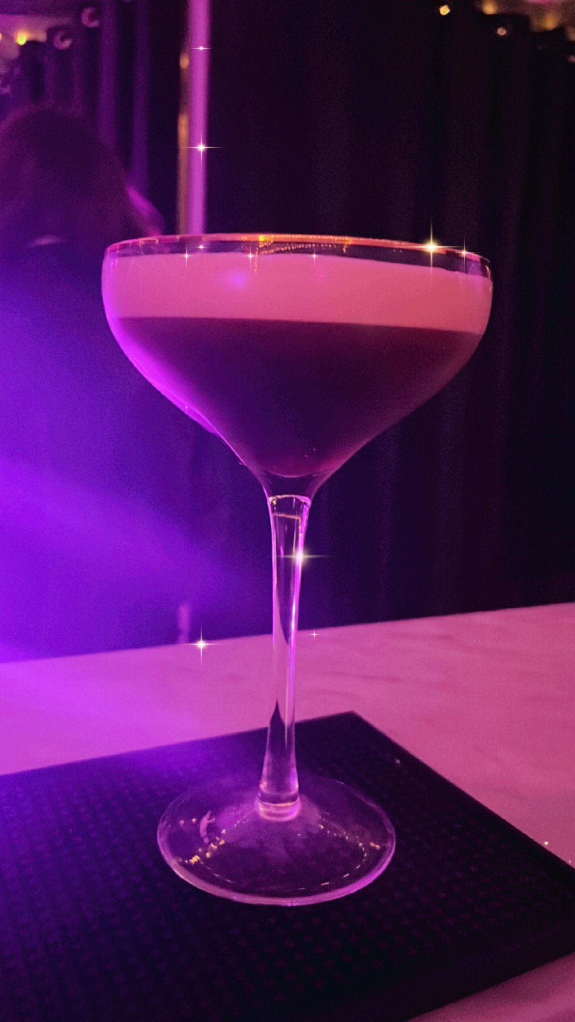 A glass of pink cocktail with a layer of foam on top, illuminated by purple and pink lights, placed on a black textured mat.
