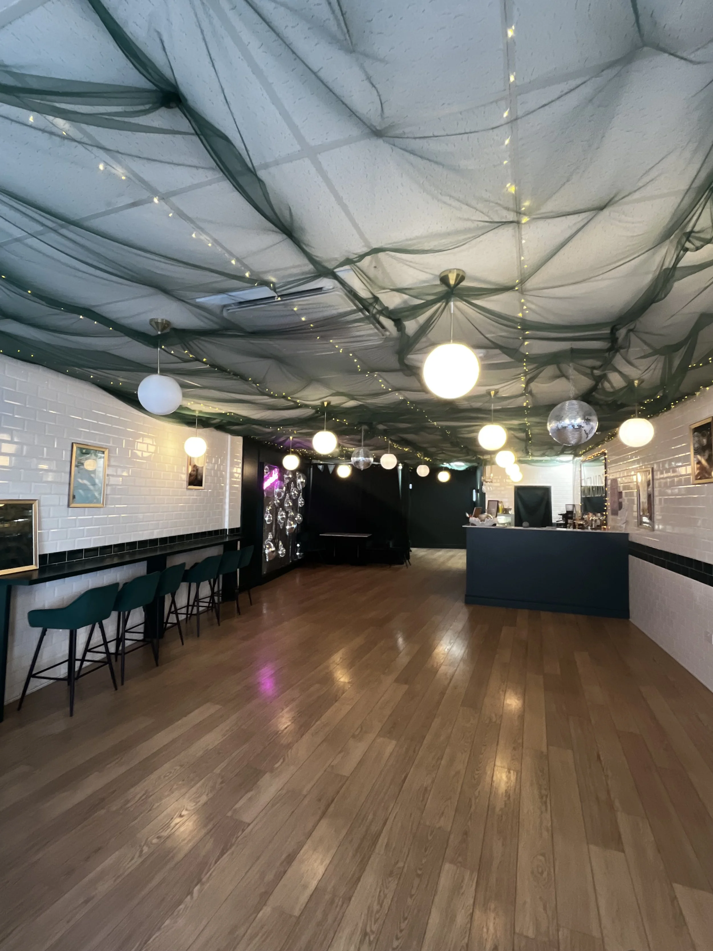 Interior of a decorated event space with a dark ceiling draped with green fabric and string lights, white brick walls adorned with framed art, a row of green chairs along a wall, and a counter on the right side.