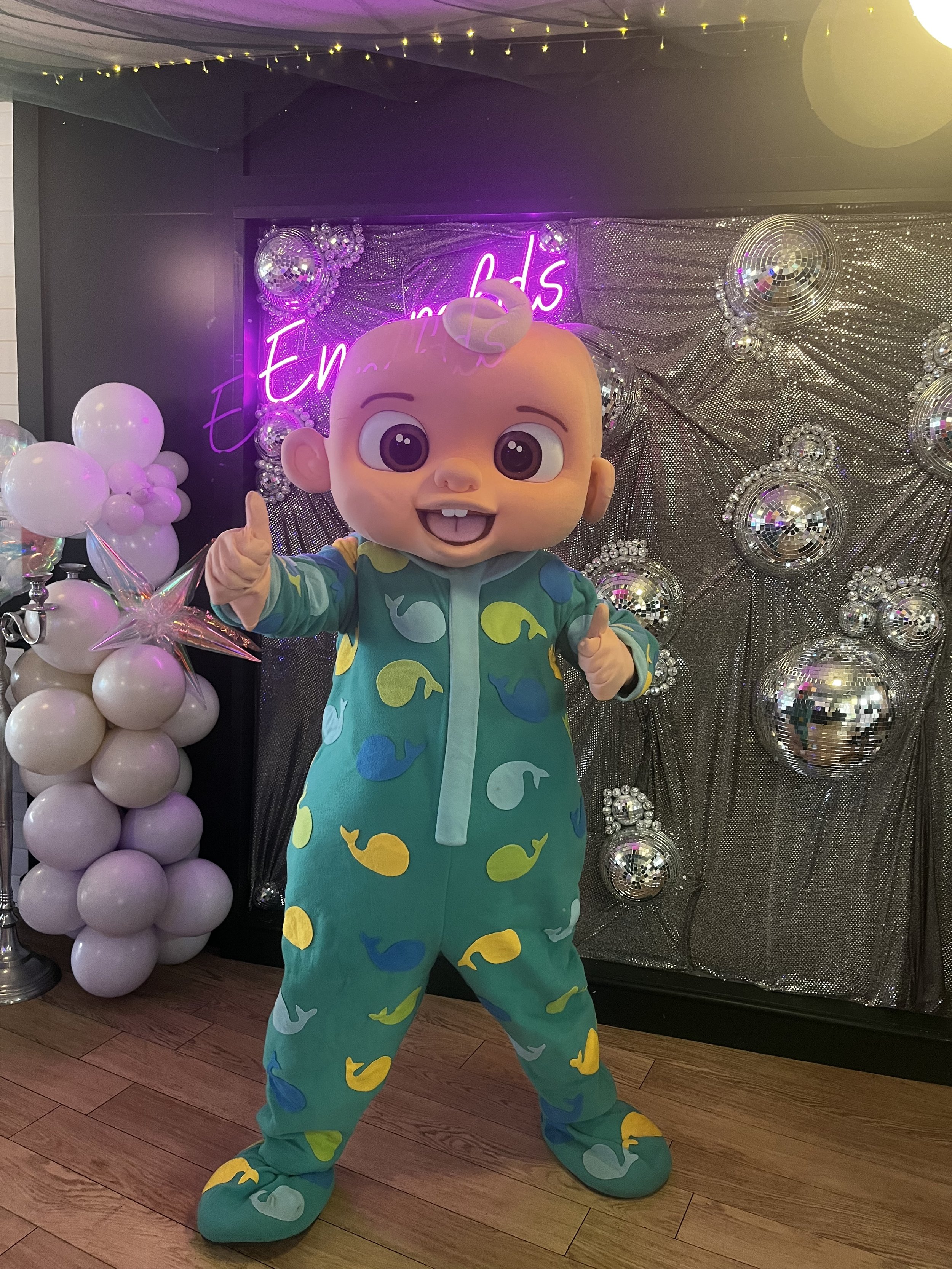 A person dressed in a large baby costume with a pacifier headpiece, wearing a colorful onesie with a dinosaur pattern, standing in front of a metallic, glittery backdrop decorated with disco balls and a neon sign that reads 'End of'.