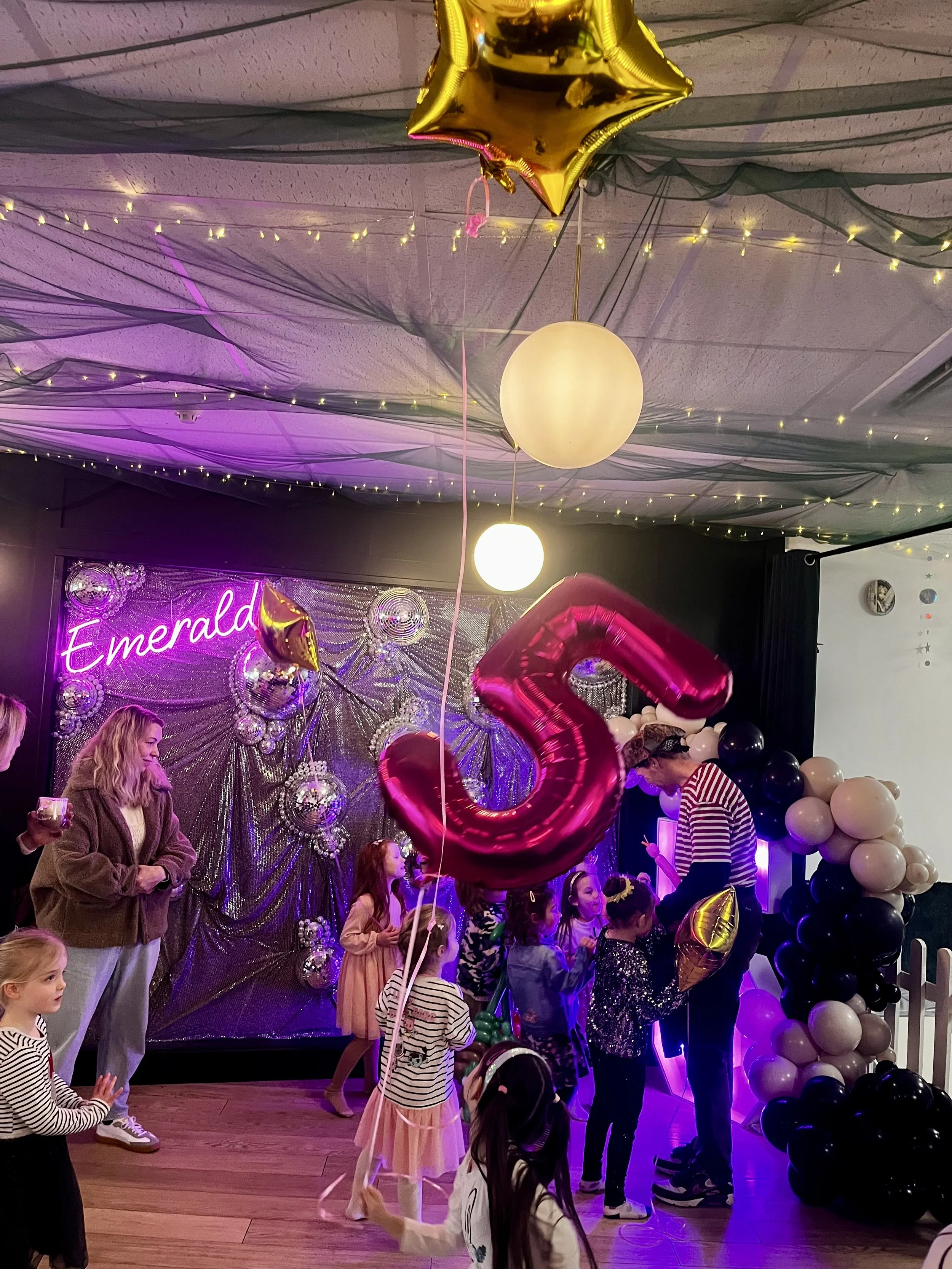 Children and adults celebrating at a party with black and white balloons, pink and gold balloons, a large pink letter 'G' balloon, and a backdrop decorated with silver and purple disco balls, a neon sign that reads 'Emerald', fairy lights, and black 