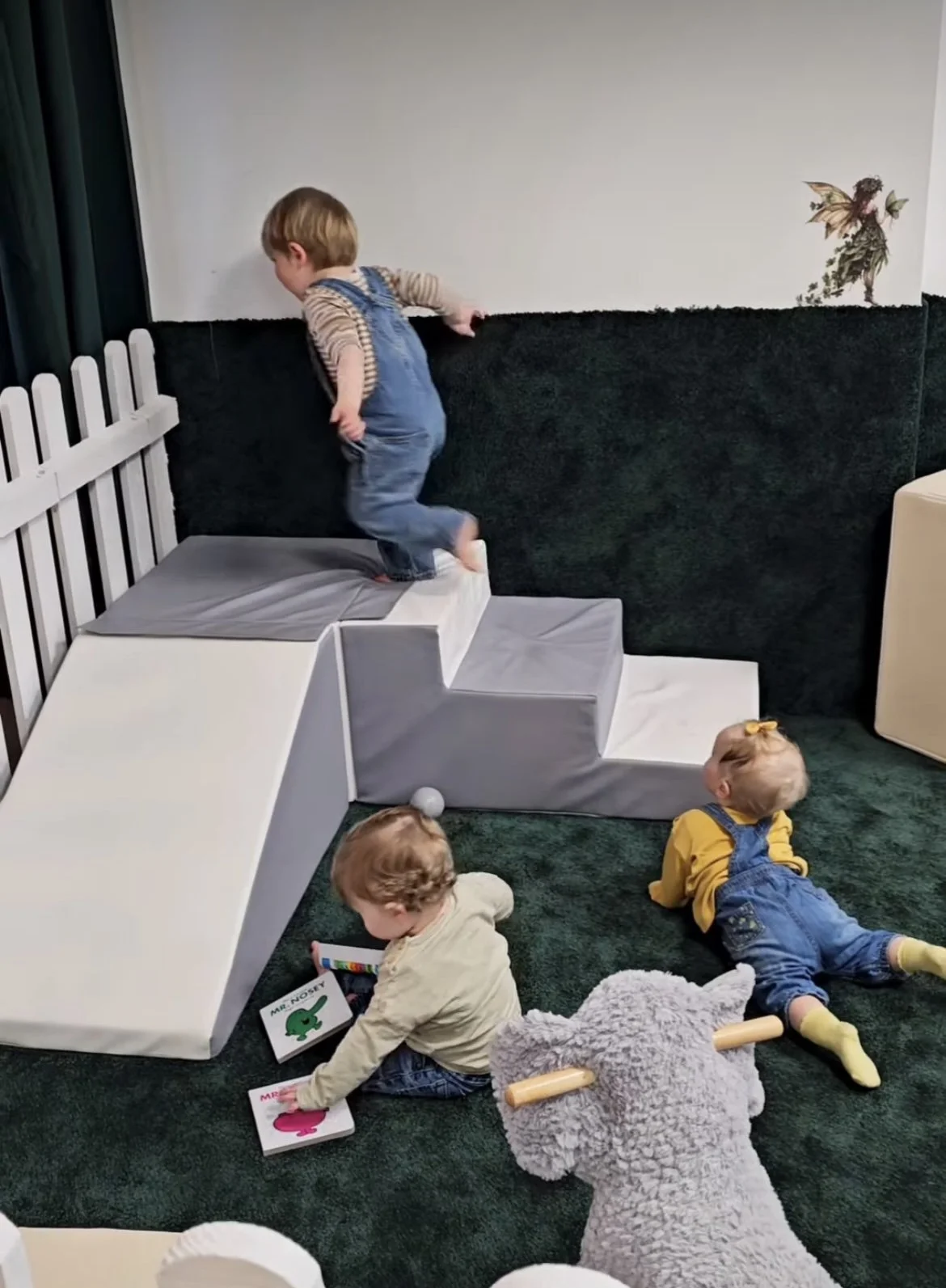 Four children playing on a green carpeted floor, with a small white and gray climbing structure and a wall decorated with a fairy sticker. One child is climbing the structure, another is sitting and reading a book labeled 'Mr. Nasty,' and two others are lying on the floor, one in a yellow shirt and the other with a stuffed animal resembling a dog.