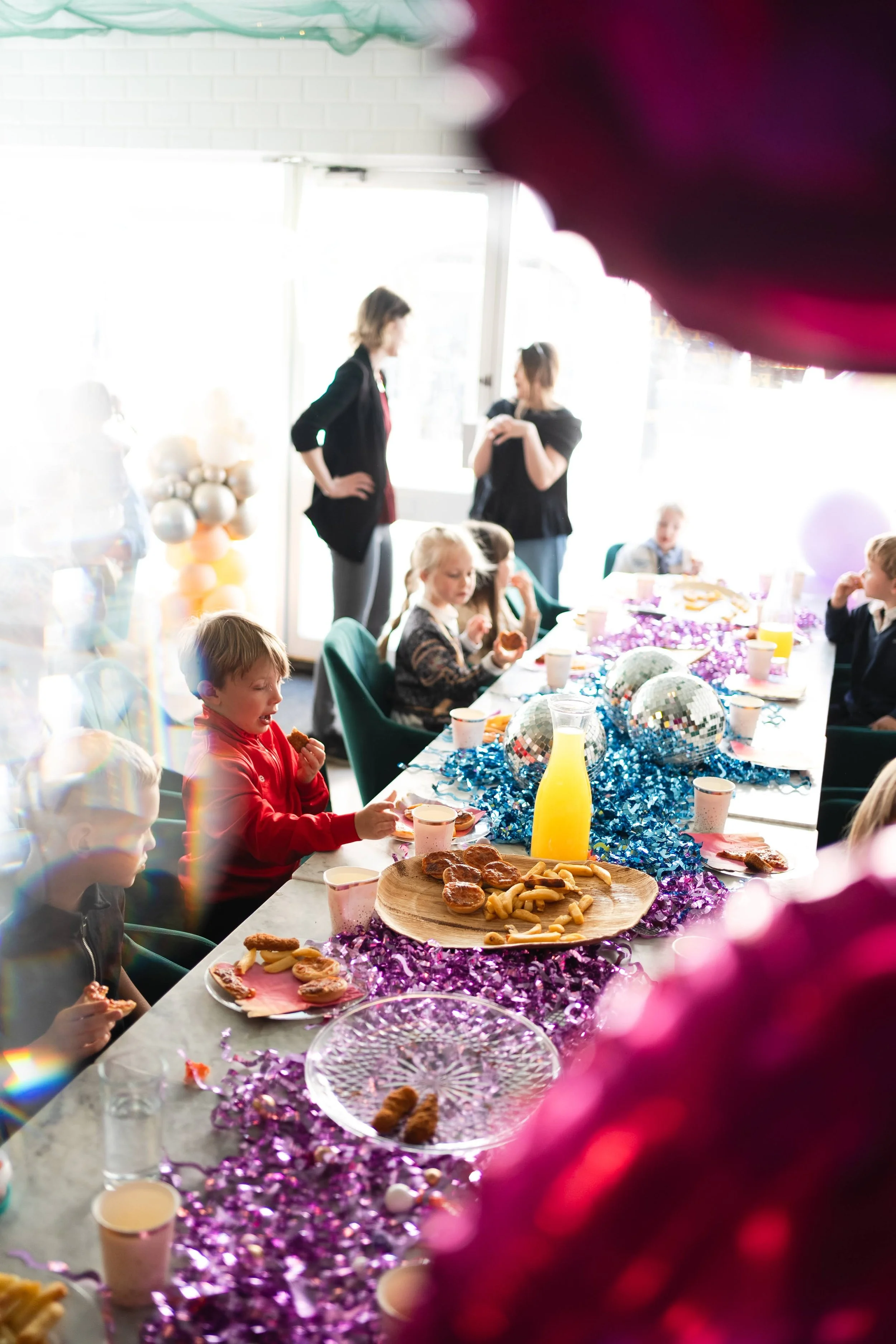 Children and adults at a party table decorated with purple tinsel and balloons, with snacks and festive treats on plates, enjoying a celebration in a bright room.