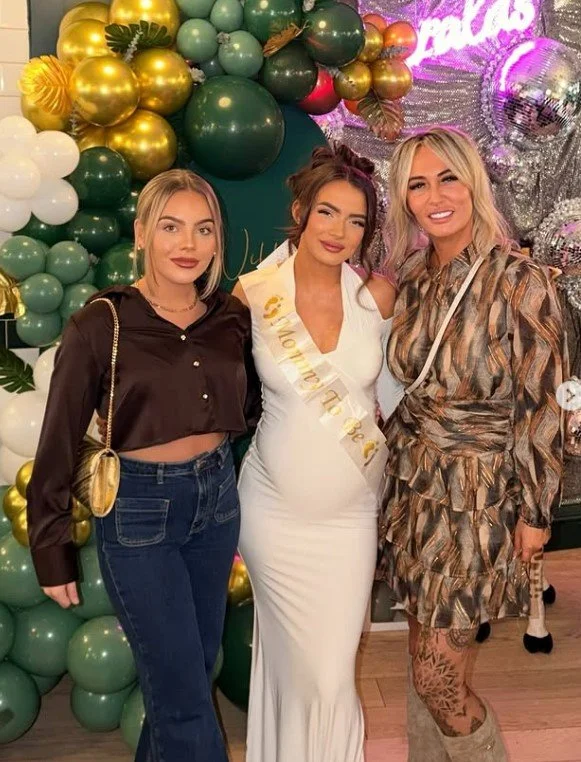 Three women standing in front of a decorated balloon background at a celebration. The woman in the middle wears a white dress with a sash that says 'Maternity', indicating a pregnancy celebration. The woman on the left wears a cropped black blouse an