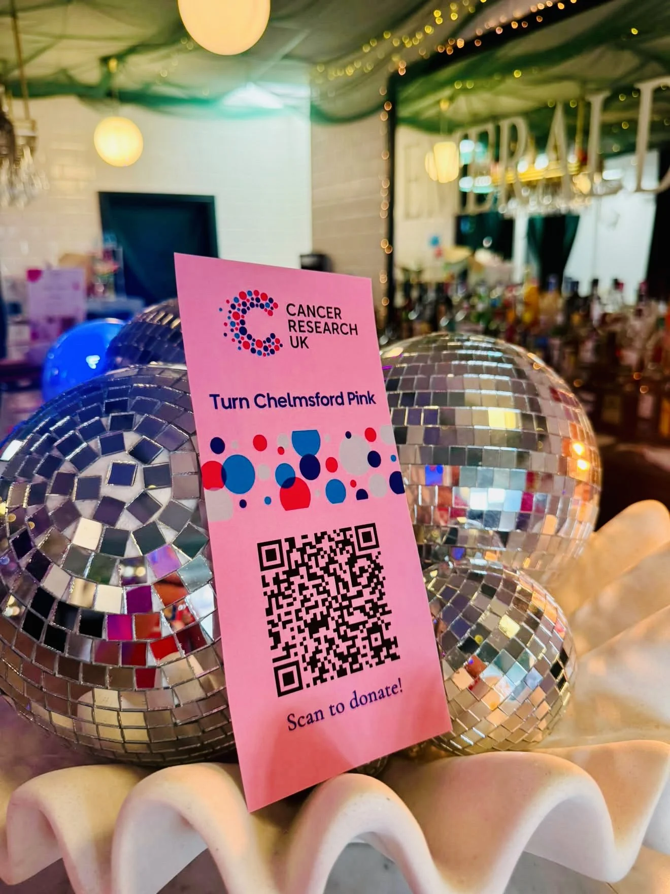 A pink sign promoting Cancer Research UK with a QR code for donations, resting on shiny disco balls, and a blurred background of a decorated event space.