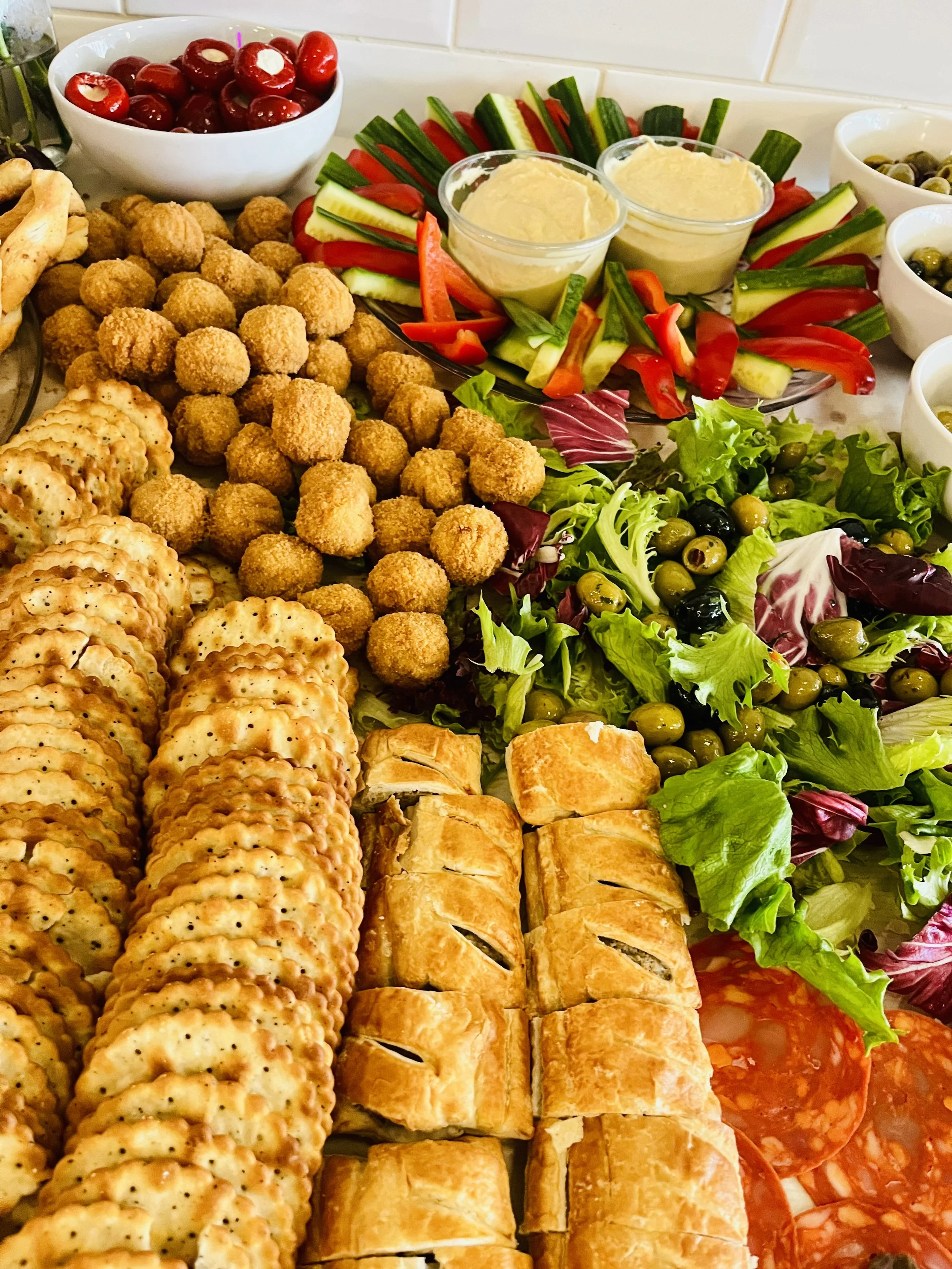 Assorted snacks including crackers, fried balls, pastries, fresh vegetables, salad, and bowls of olives and cherries.