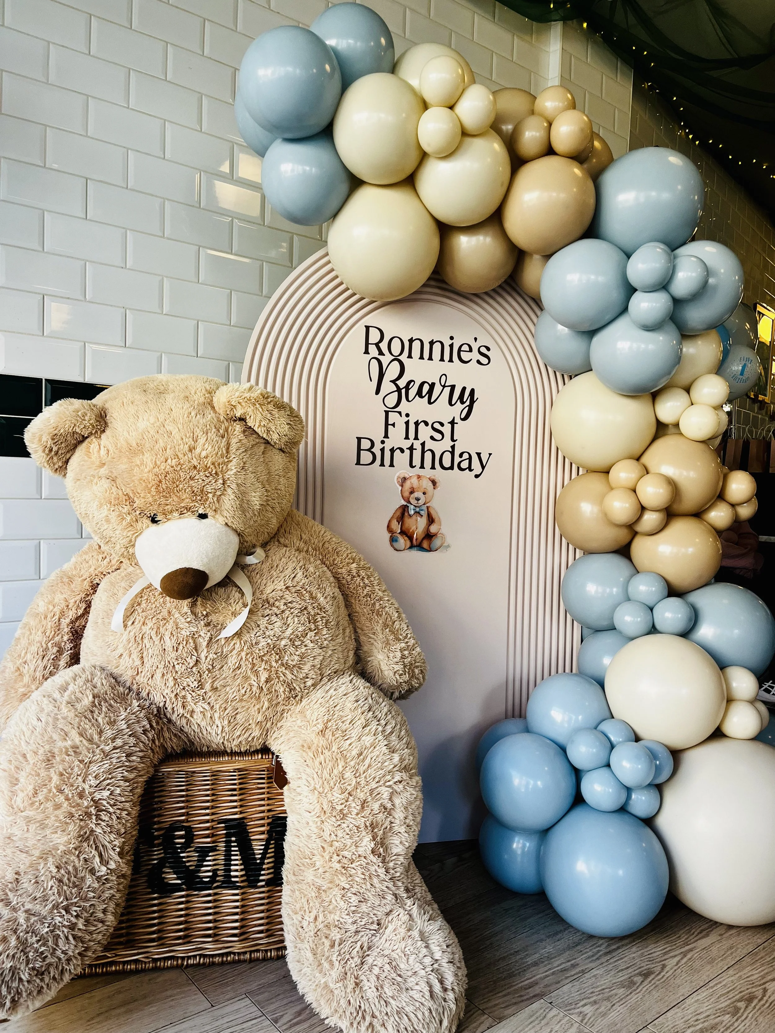 Large teddy bear sitting on a woven basket with a mini teddy bear picture on it, in front of a backdrop with pastel-colored balloons and a sign that reads 'Ronnie's Beary First Birthday'.