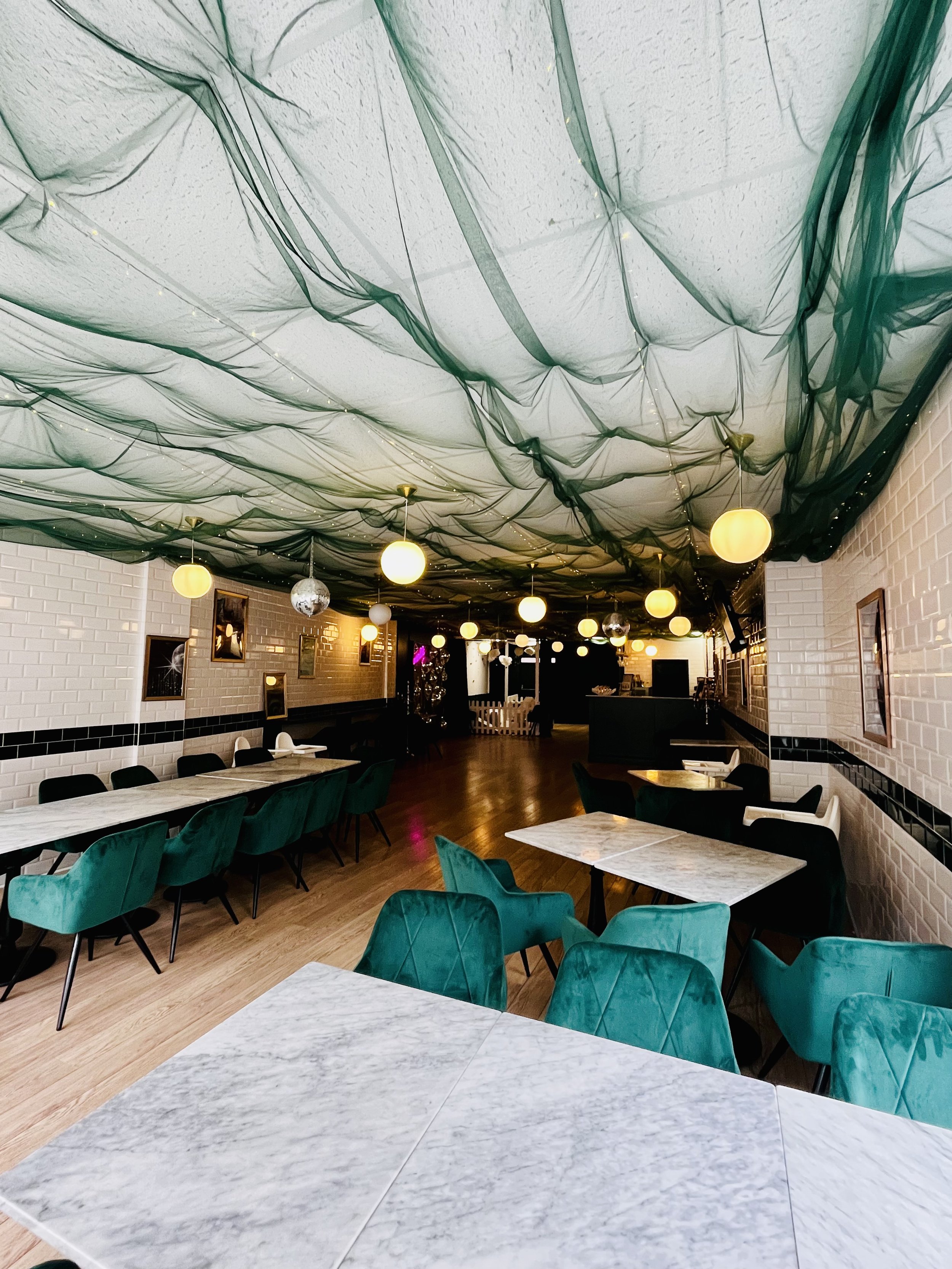Interior of a decorated restaurant or cafe with fabric draped ceiling, string lights, round pendant lights, and tables with green velvet chairs and marble tabletops.