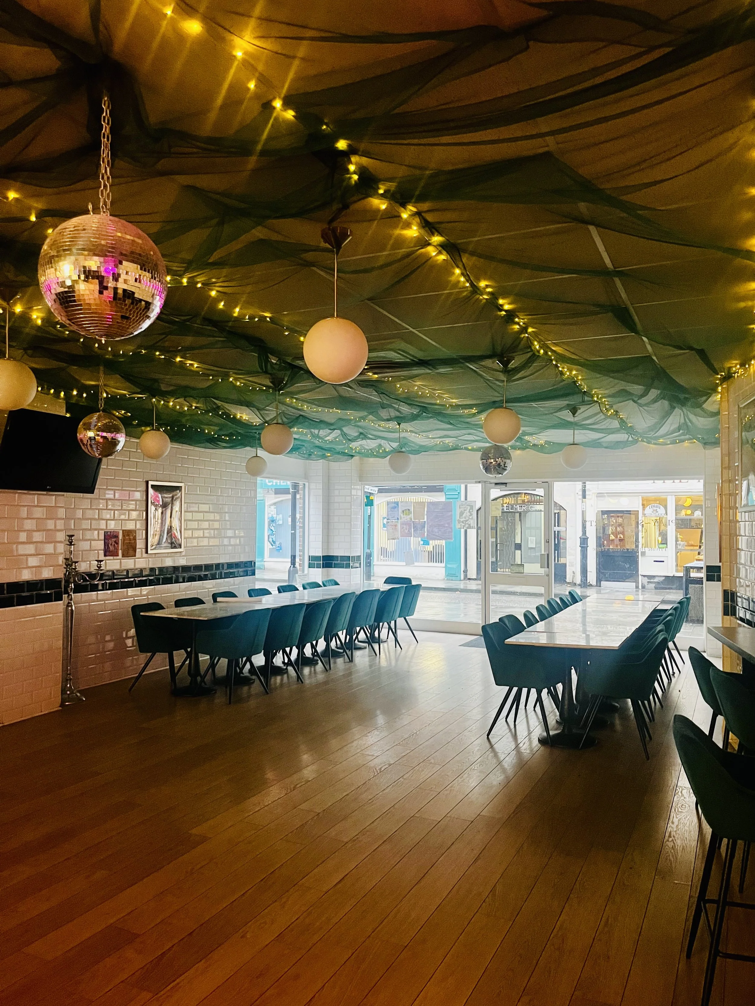 Interior of a restaurant with wooden flooring, dark green chairs arranged around tables, brick walls, large windows at the front, and a ceiling decorated with black fabric, string lights, and disco balls.