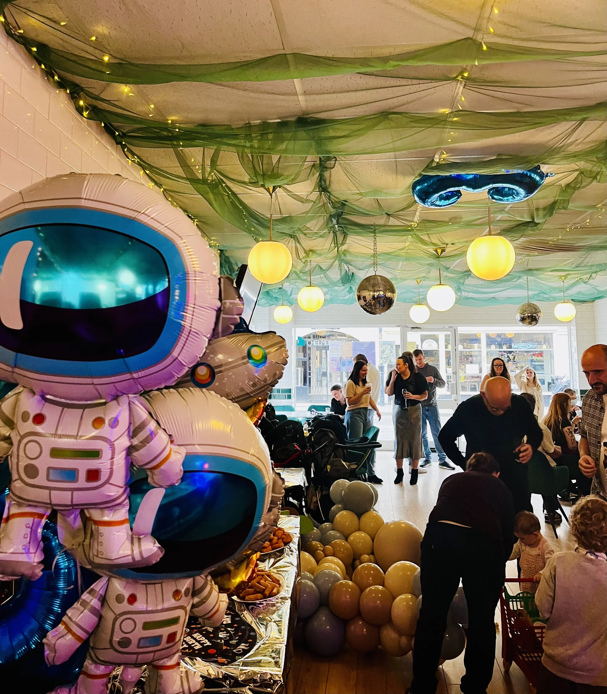 Indoor party scene with green fabric drapes and string lights on the ceiling. People are gathered around tables, some standing and chatting, others seated. Balloons in pink, purple, yellow, and silver are visible, including a large space-themed ballo