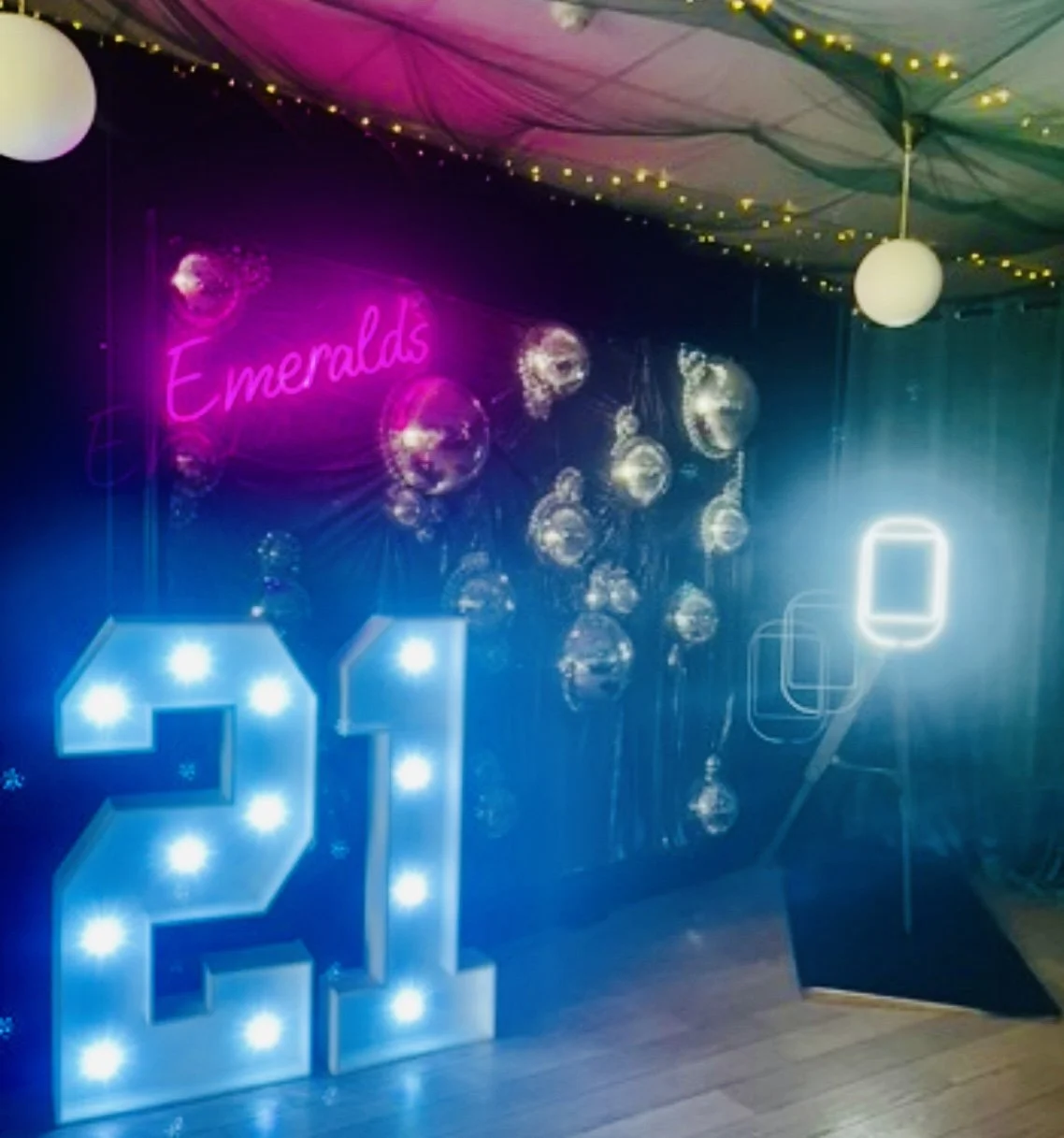 Decorations for a celebration with a neon pink sign reading 'Emeralds', large illuminated number '21', hanging balloons, and string lights on a decorated ceiling.