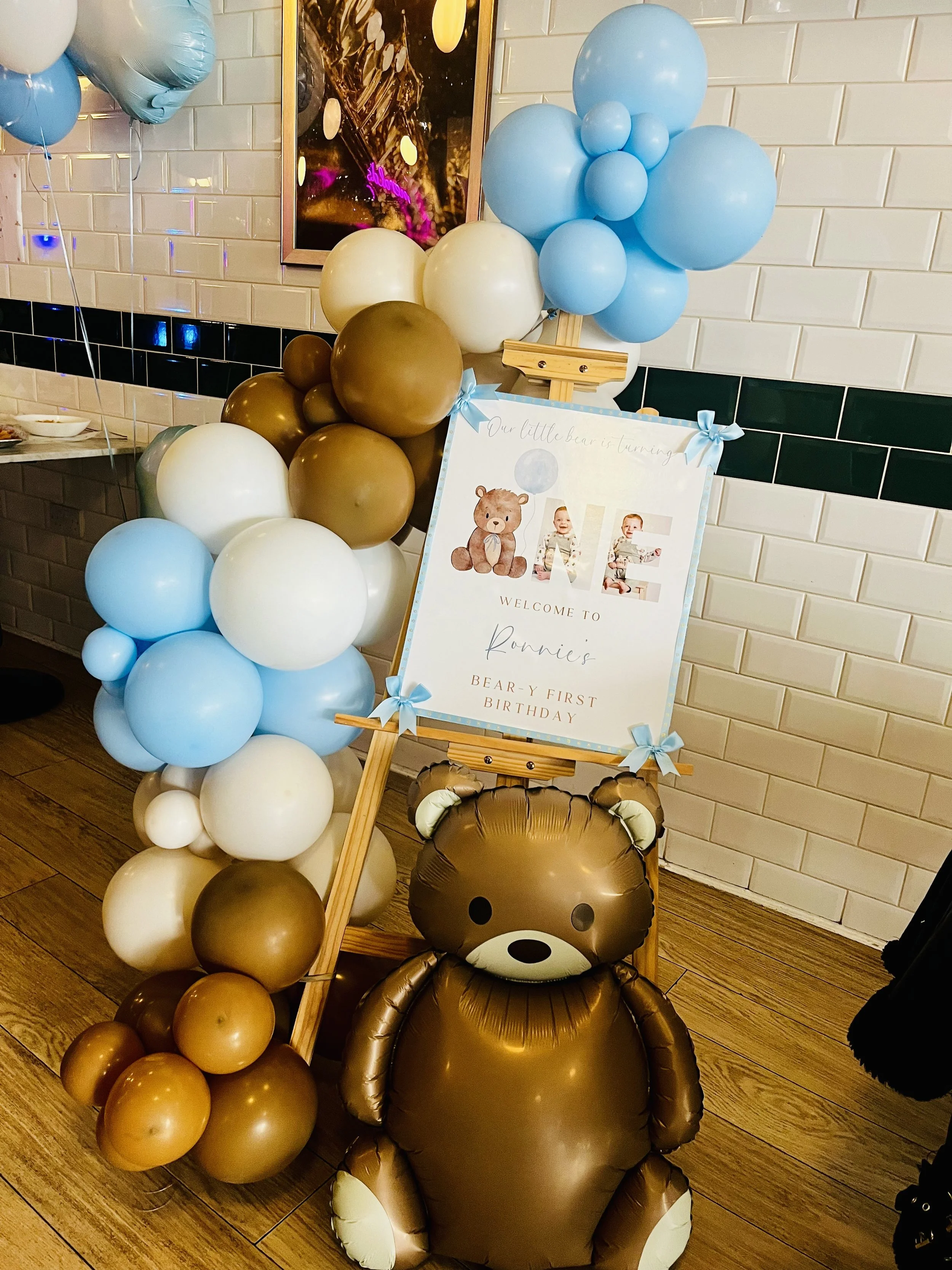 Decorations for a child's first birthday, featuring balloons in blue, white, and brown arranged around a wooden easel with a sign that has images of a bear and children, and text that reads 'Our little bear is turning' and 'Ronnie's bear-y first birt