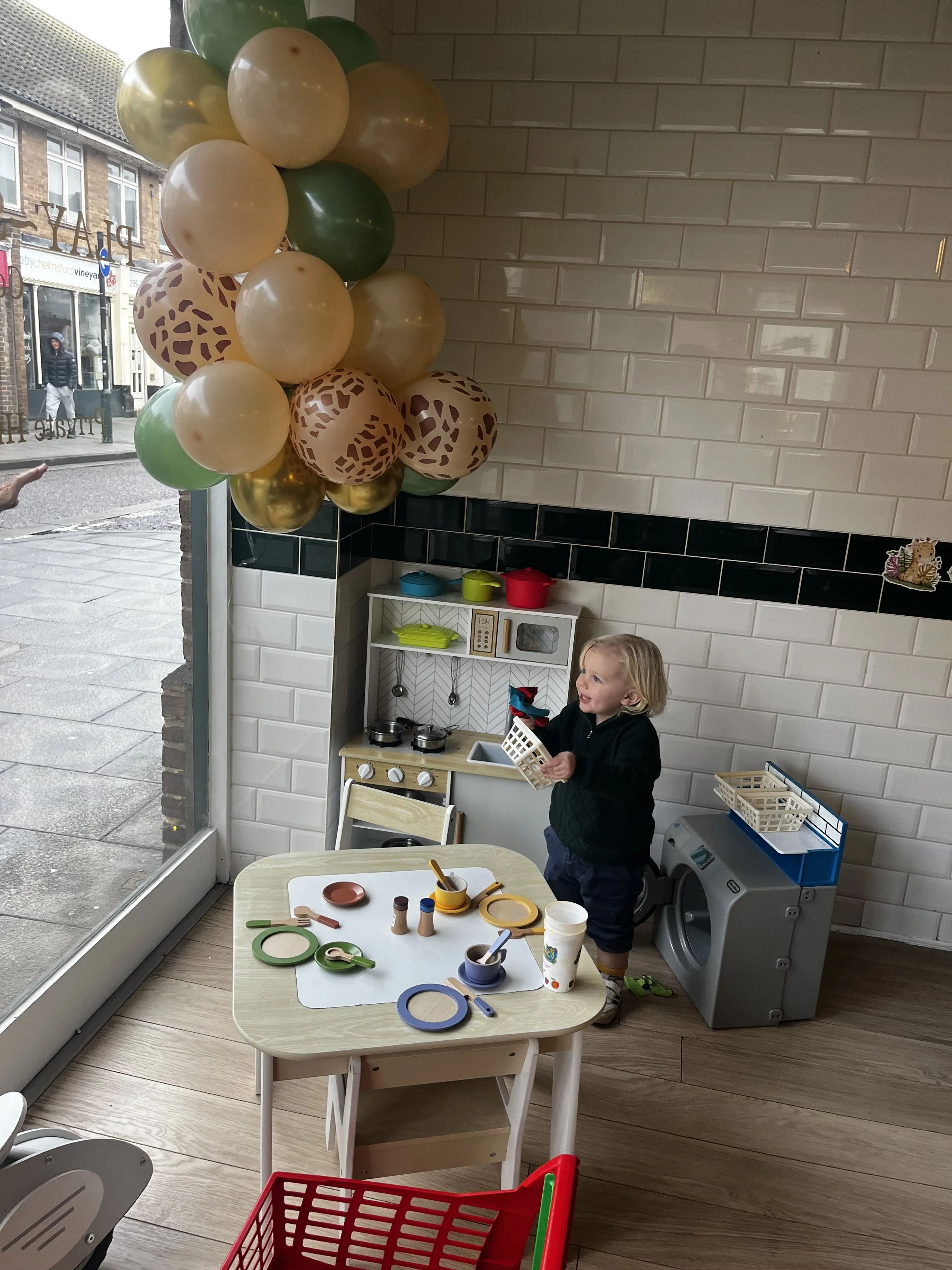 A young boy in a play kitchen area inside a cafe or bakery, playing with toy food and kitchen utensils, with balloons outside the window.