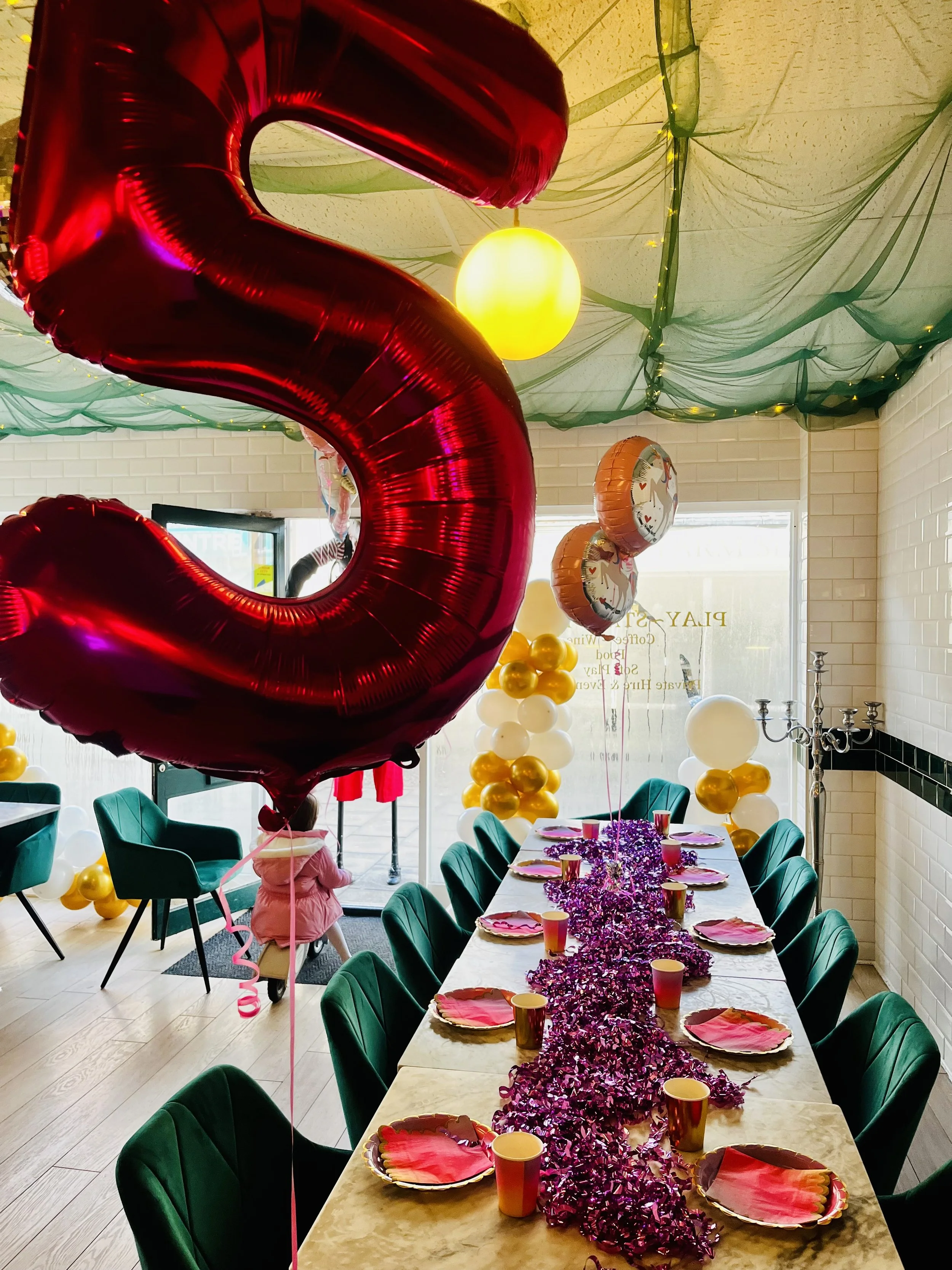 Party room decorated with balloons, a long table with pink and yellow disposable cups, pink napkins, and pink tinsel table runner, surrounded by green chairs, with balloons and a large red number 2 foil balloon in the foreground, green fabric draping