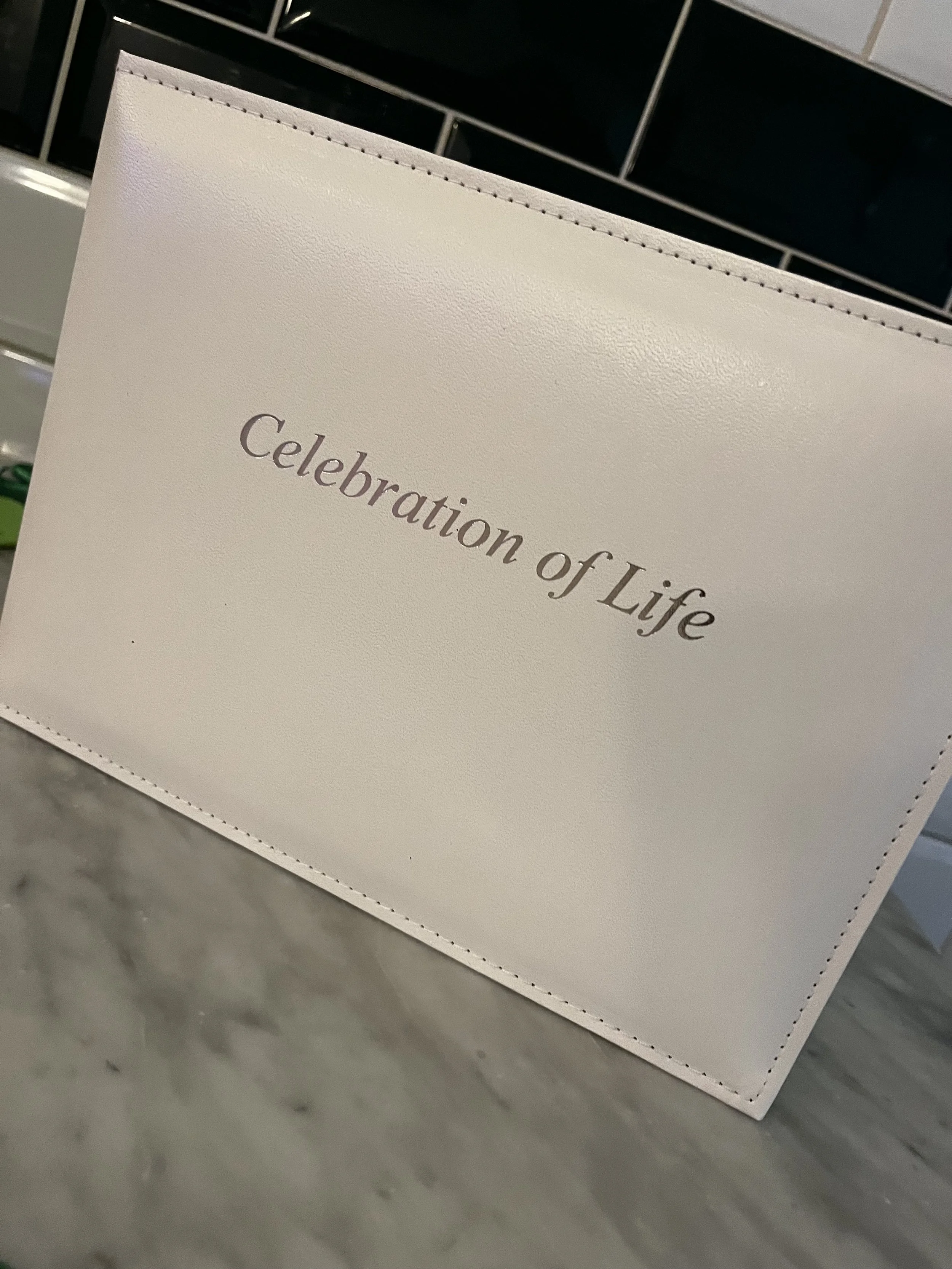 A white card or sign with the words 'Celebration of Life' written on it, placed on a marble surface with tiled wall in the background at emeralds chelmsford